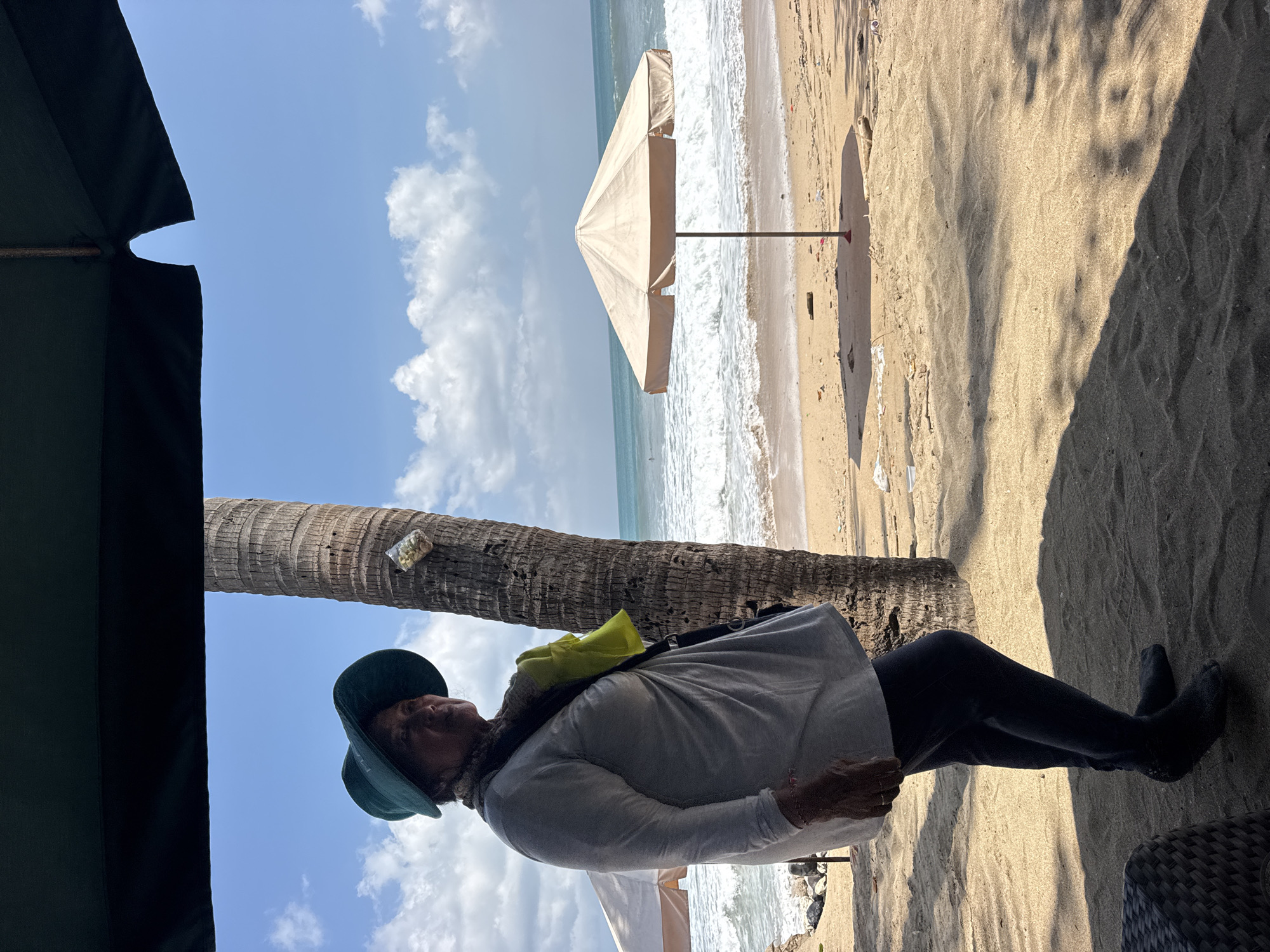 A woman wearing a green hat, a grey long-sleeved shirt, and dark pants stands on a sandy beach in Kuta, Indonesia. She has a yellow bag slung over her shoulder. She is standing next to a palm tree, and a beach umbrella is visible in the background. The ocean waves are crashing on the shore. The sky is blue with scattered clouds. The image appears to be taken from under a dark canopy, possibly an awning or a large umbrella. The lighting suggests it is daytime, with sunlight casting shadows on the sand.