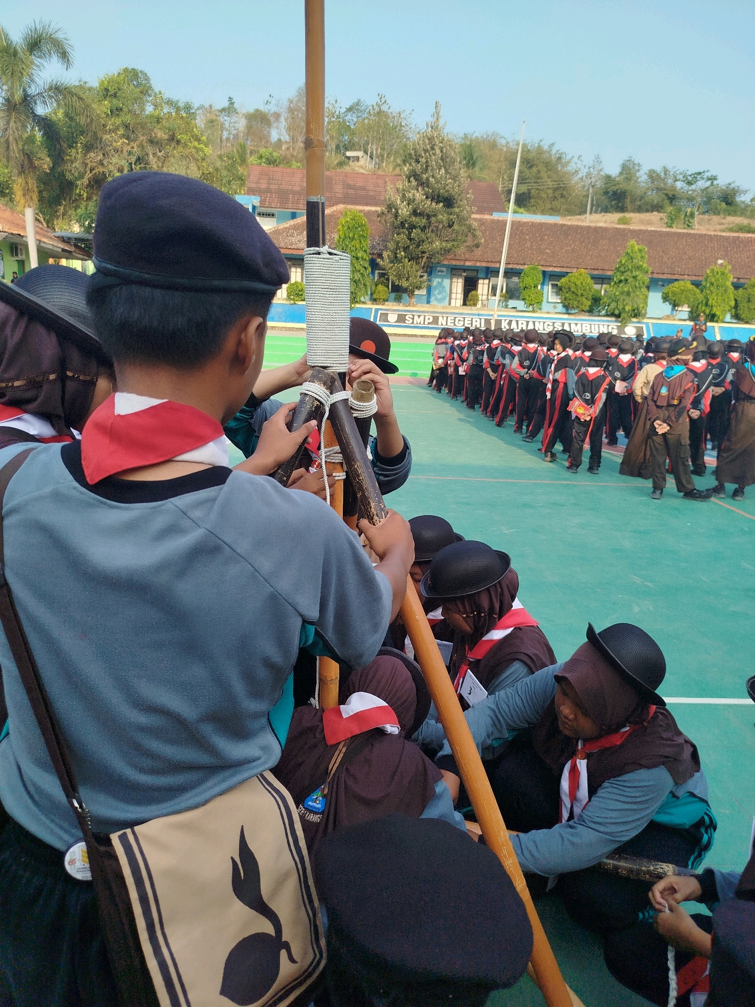 In Karangsambung, Indonesia, a group of young scouts, likely students from SMP Negeri 1 Karangsambung, are gathered on a teal-colored sports field. They are engaged in an activity involving a wooden flagpole. The scouts are wearing uniforms consisting of dark blue berets, grey long-sleeved shirts, dark pants, and red and white neckerchiefs. Some are wearing brown vests over their shirts. Several individuals are focused on tying ropes to the flagpole, indicating a camping or flag-raising ceremony. In the background, a school building with brown tiled roofs and blue walls is visible, along with trees and a clear sky. The overall atmosphere appears to be a formal gathering or training exercise.