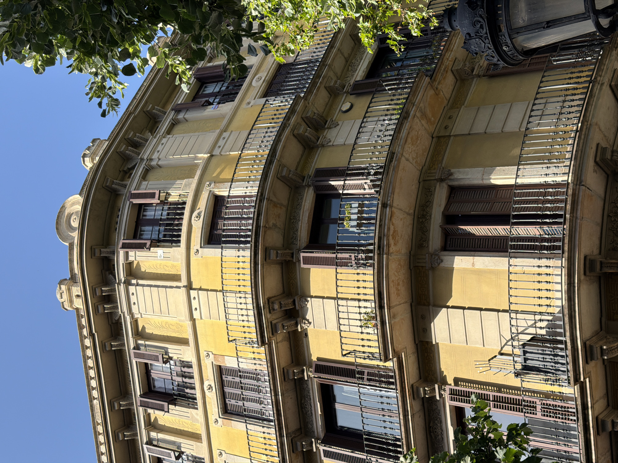 A close-up, low-angle shot captures a section of a yellow, ornate building facade in Barcelona, Spain, under a clear blue sky. The building features multiple stories with curved balconies, each adorned with black wrought-iron railings. The balconies are supported by decorative stone corbels. Dark brown shutters are open on several windows. Some of the windows show faint reflections or interior details. Lush green foliage from a tree partially obscures the upper right portion of the building. In the lower right corner, the top of a black, antique-style lamppost with ornate detailing is visible. The architectural style suggests an older, possibly Art Nouveau or similar period construction.