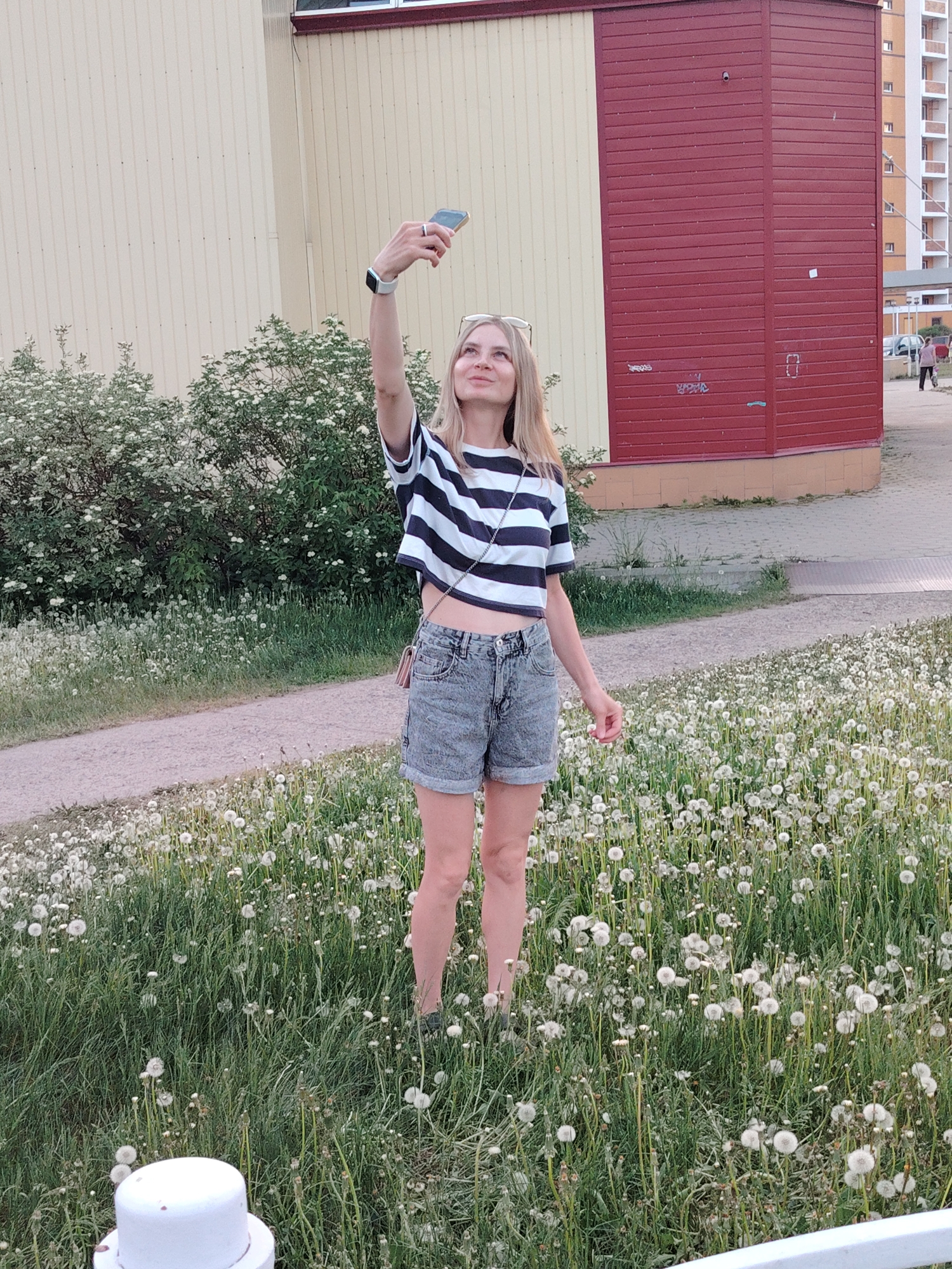 A fair-skinned, blonde woman in a striped, cropped shirt and denim shorts takes a selfie in a grassy field of dandelions. She holds her phone aloft in her right hand, wears a smartwatch, and gazes upwards. The location appears to be an urban area, specifically Chaikovsky, Russia, as indicated by architectural styles. Buildings with colored facades stand in the background, as well as a paved pathway and green bushes. Another person is walking in the background.