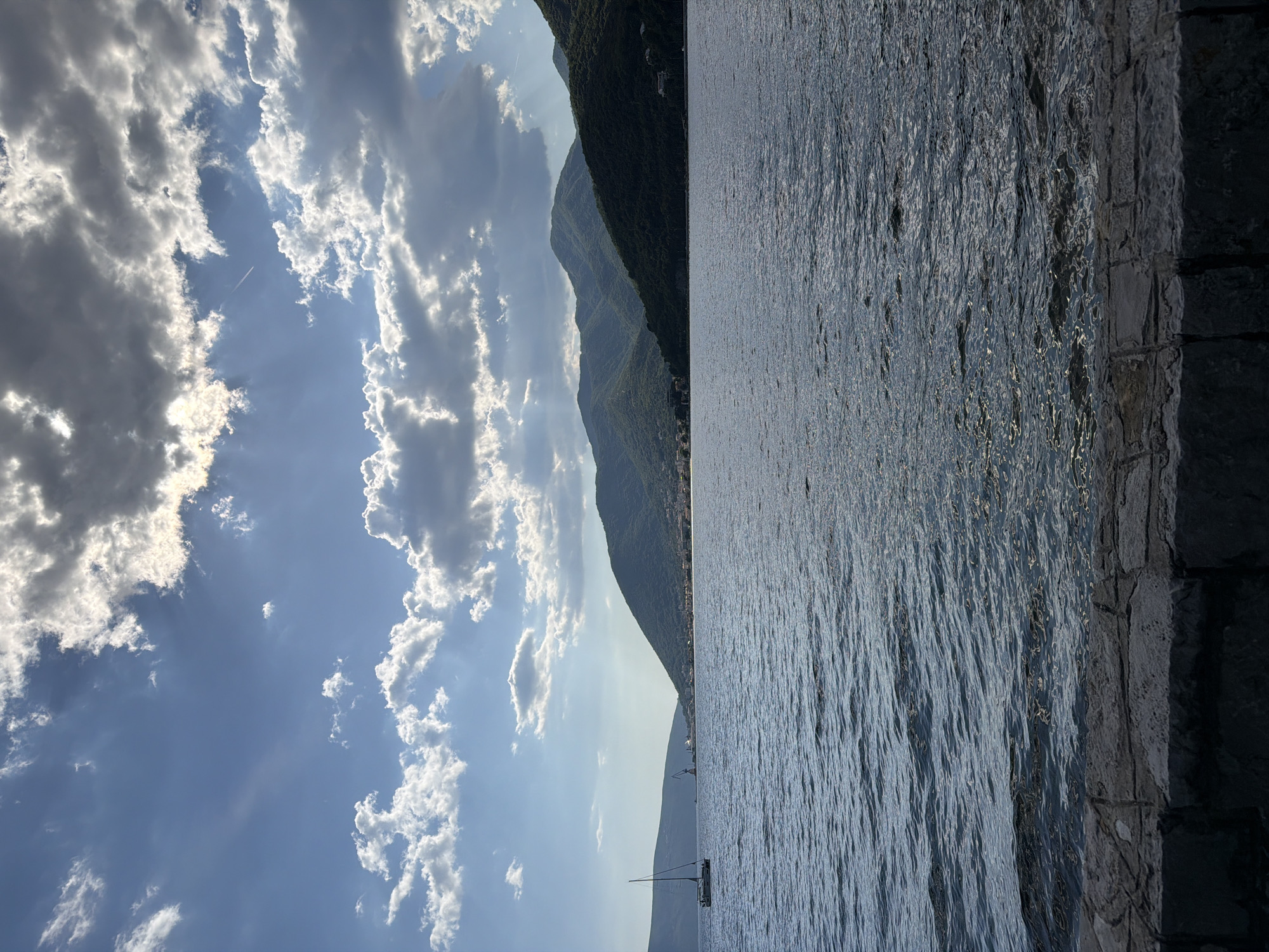 The image depicts a landscape with a body of water, mountains, and sky. On the right is a stone structure or wall. The water is calm, with reflections of light on the surface. In the distance, there are green mountains. Above, the sky is blue with scattered white clouds. A small sailboat is visible on the water. The location is Donja Lastva, Montenegro.