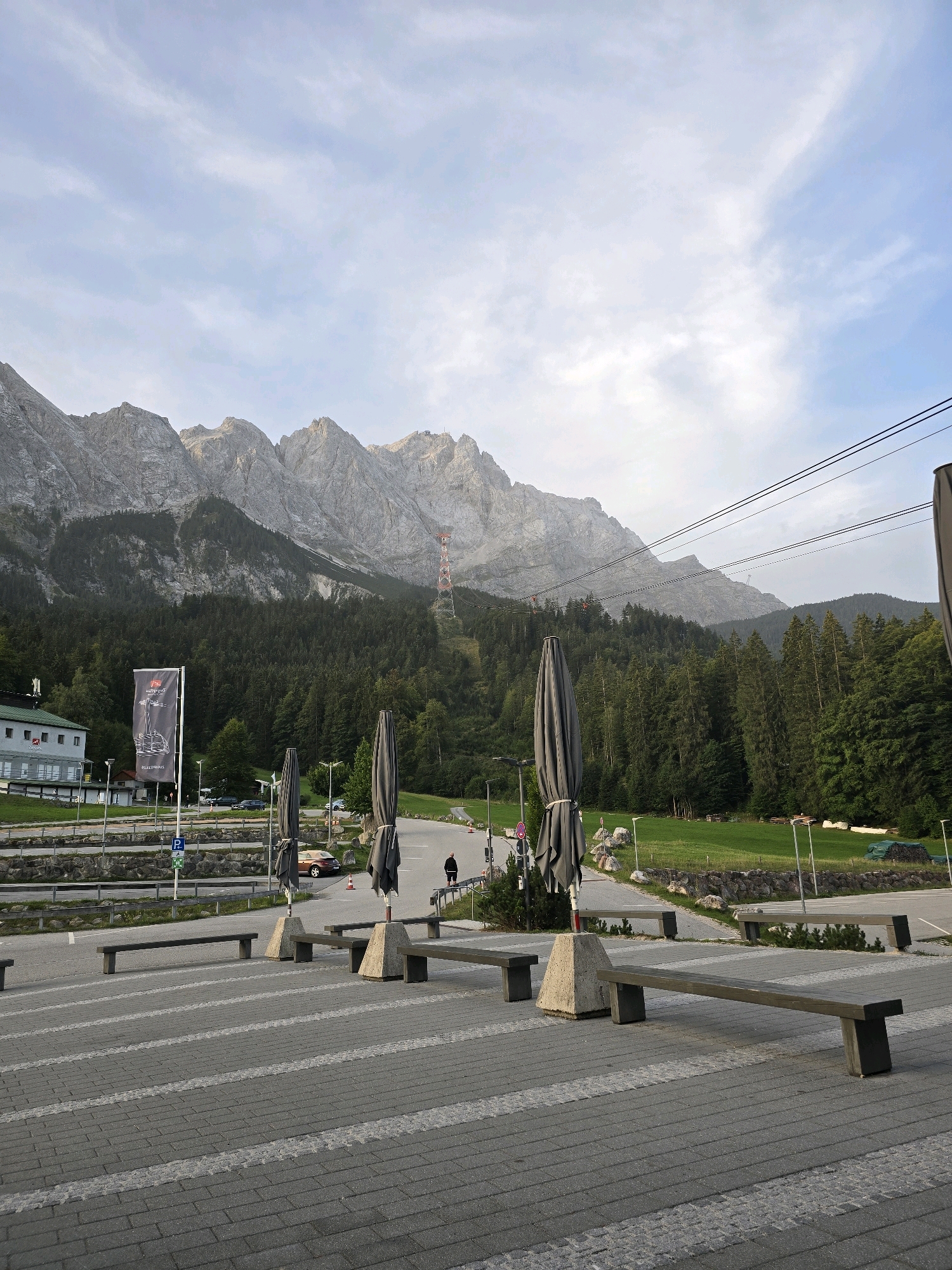 This is a full-shot, eye-level, outdoor photograph taken in Grainau, Germany, likely at the base of the Zugspitze mountain. The scene depicts a paved area in the foreground with several long, dark benches supported by concrete blocks. Two large, closed gray umbrellas stand on either side of the paved area.
In the mid-ground, a road curves to the right, with several cars parked and one person walking. Beyond the road is a grassy area and a dense forest. In the background, a large, rugged mountain range dominates the horizon, with a ski lift tower visible on its slopes. A building with a green roof and light-colored walls is situated to the left of the mountain. A tall flag with a dark banner hangs on a pole in front of the building. The sky is overcast with scattered clouds.
