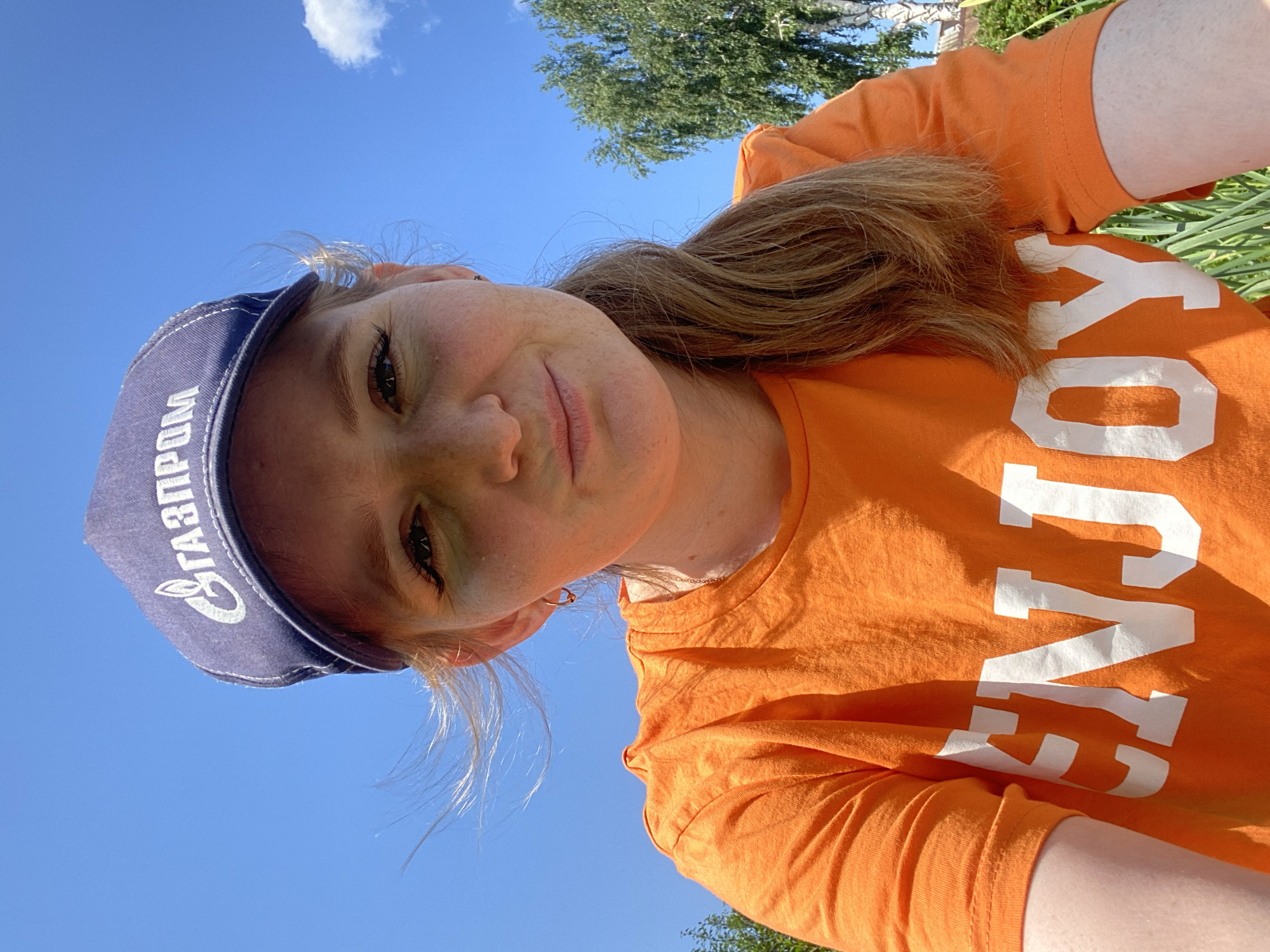 This is a vertically oriented, close-up selfie of a young woman outdoors. She is wearing a blue baseball cap with white lettering and an orange t-shirt with large white lettering. Her hair is blonde and pulled back. She is looking at the camera with a slight smile and her face is freckled. The background is a bright blue sky with a single white cloud. Green trees and foliage are visible in the upper right portion of the image. The sun appears to be shining, suggesting it is daytime. The visible text on the cap reads "ГАЗПРОМ", which is the name of a Russian energy company. The text on her t-shirt is partially visible, with the letters "JOY" or "OY" clearly discernible. The scene suggests a casual, outdoor setting, possibly in Chaikovsky, Russia, given the company logo.