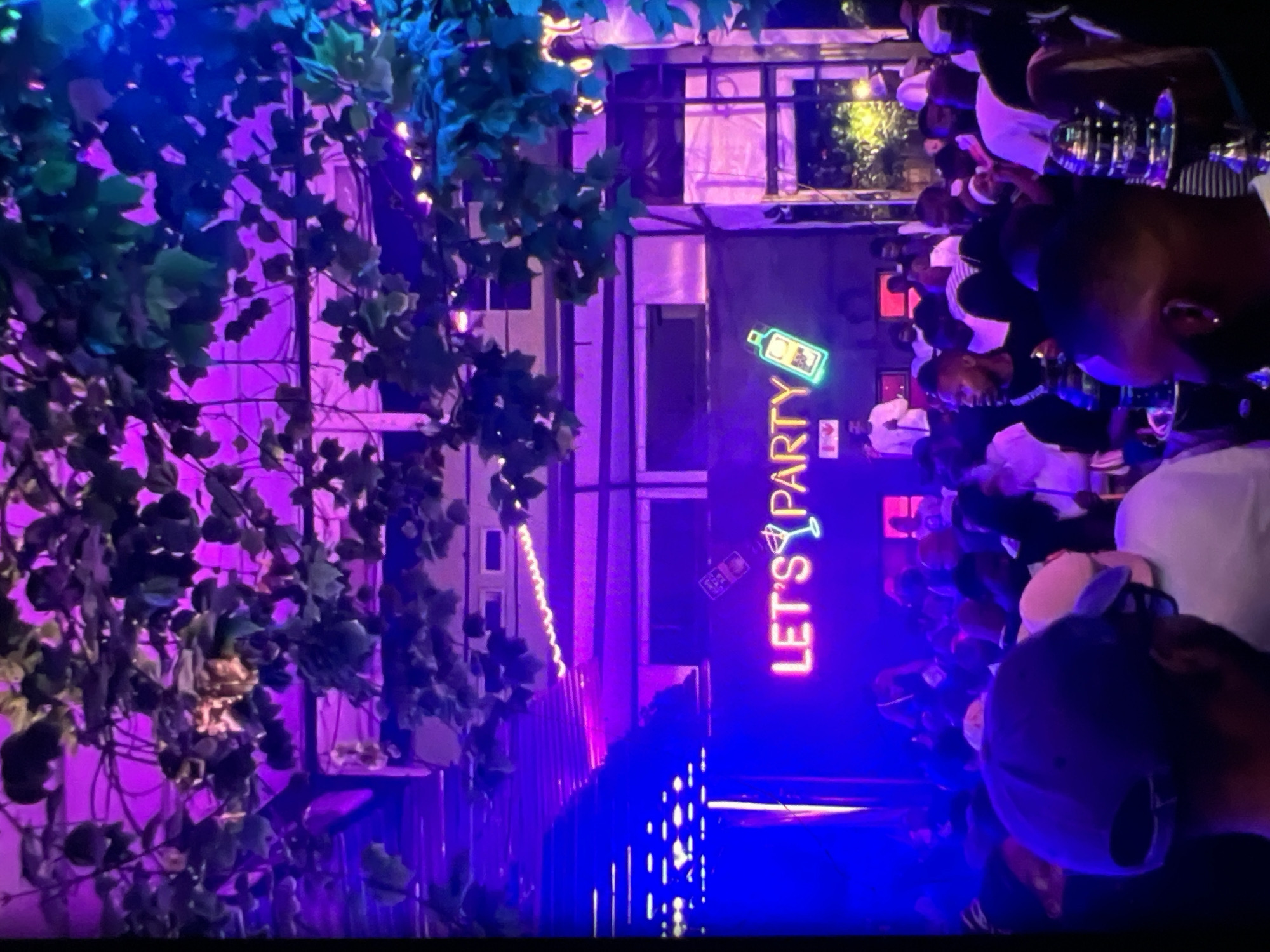 The image depicts a vibrant nightlife scene at a venue in Lagos, Nigeria, illuminated by abundant neon lights and ambient purple and blue hues. A prominent neon sign reads "LET'S PARTY" in bright pink and features a martini glass and a bottle graphic. The venue appears to be an outdoor or semi-outdoor space, with overhead foliage and structures suggestive of a rooftop bar or a similarly open-air establishment. A diverse crowd of people are gathered, many appearing to be enjoying themselves, with some interacting and others focused on the activities. The atmosphere is energetic and celebratory. The image focuses on the illuminated sign and the surrounding crowd.