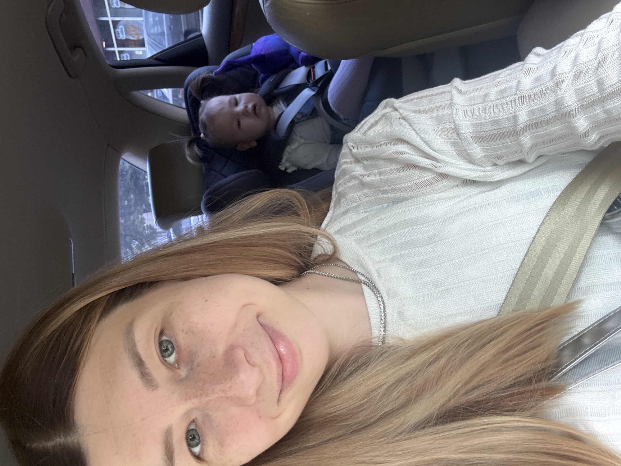 A person with light skin, freckles, and blonde hair is taking a selfie. The person is wearing a white shirt and a silver necklace. A child in a car seat is visible in the background. The child appears to be sleeping. The location cannot be determined from the image.