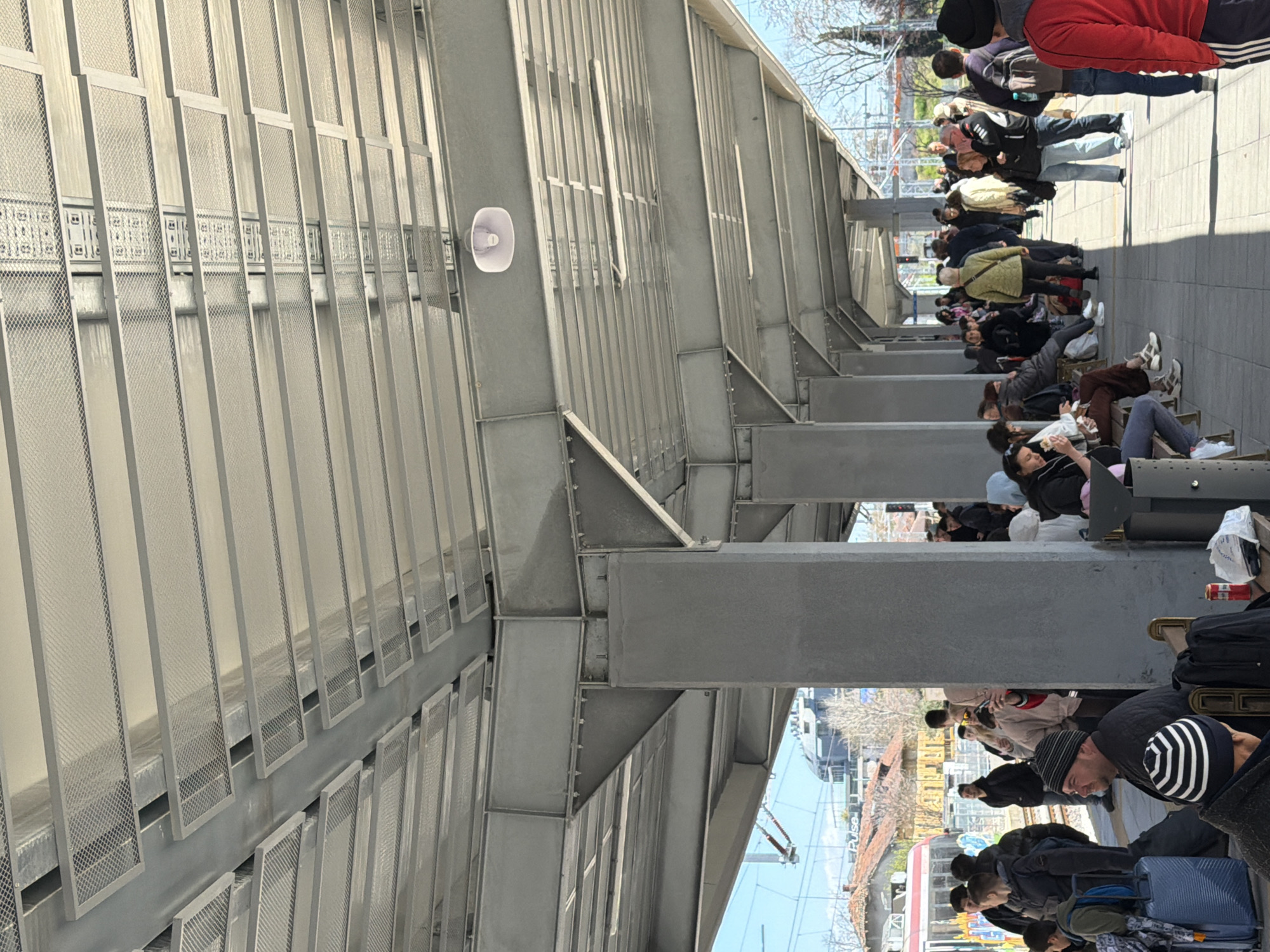 The image depicts a group of people waiting at what appears to be a train or bus station platform in Plovdiv, Bulgaria. The people are lined up along a covered walkway or waiting area, some sitting on benches and others standing. Many have luggage with them, including suitcases and backpacks. The structure providing cover consists of a gray, metallic framework with mesh-like panels forming the walls. A white loudspeaker is attached to one of the structural supports. In the background, through the structural elements, parts of the city are visible, including a train, buildings, and trees. The lighting suggests it is daytime with clear skies.