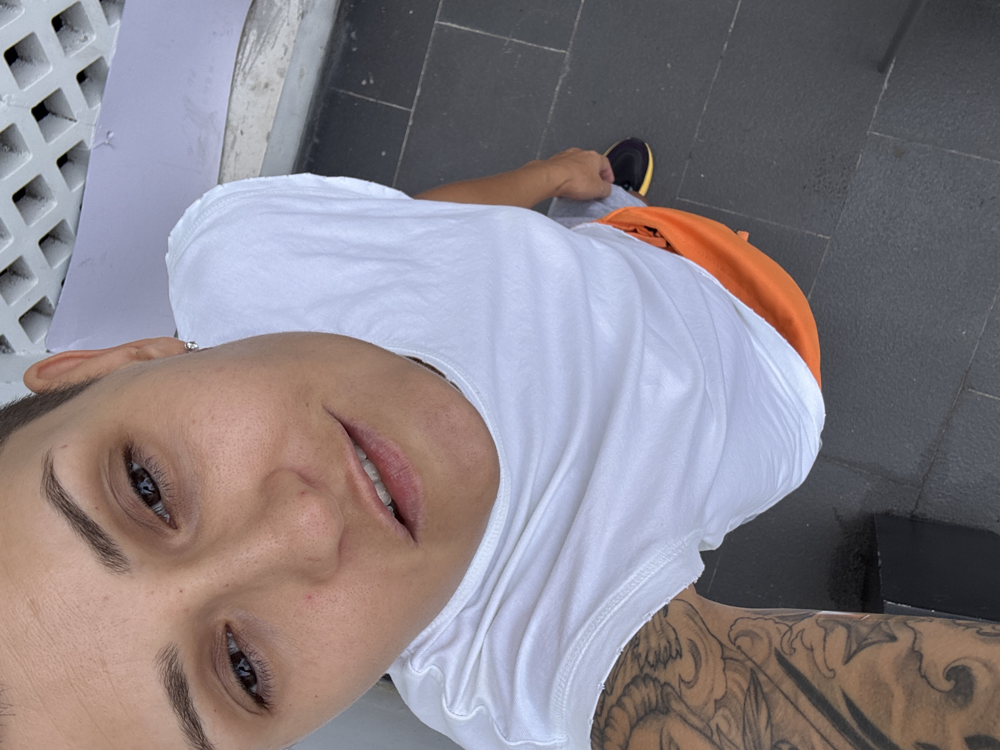 A high-angle medium shot shows a person with short hair, wearing a white sleeveless shirt and orange shorts, standing on a dark tiled surface. The person's face is turned towards the camera, with their mouth slightly open and a hint of a smile. Their left arm, heavily tattooed, is visible on the left side of the frame. In the background, a white textured wall with a grid pattern and a light purple surface are visible. The person's right foot, wearing a black and yellow shoe, is positioned on the tiled ground.
