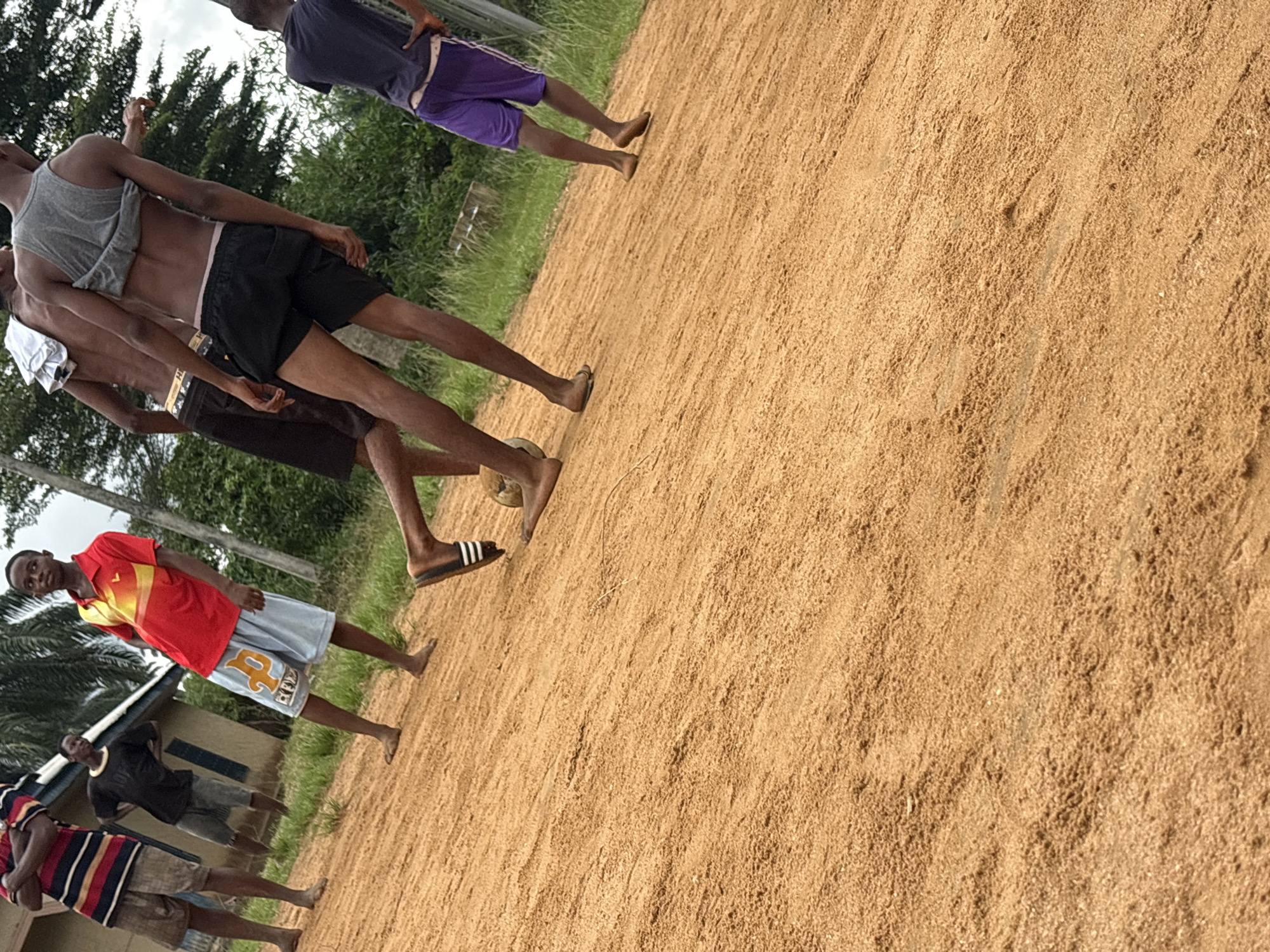 In Ikot Ekpene, Nigeria, a group of young men stand on a large sand-covered area, possibly a field or open space. One man wears a gray tank top and black shorts, another a red shirt and light blue shorts, and a third wears a striped shirt and brown shorts. Some are barefoot, and one wears Adidas slides. The background shows green vegetation and a building.