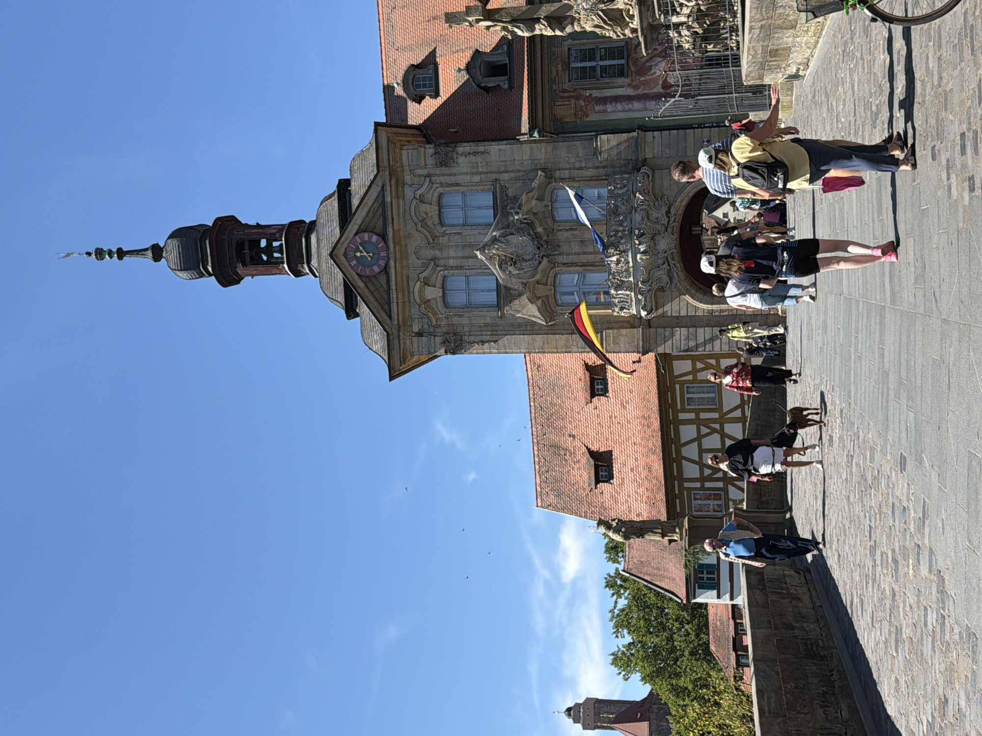 The image depicts a sunny day scene on a cobblestone bridge in Bamberg, Germany, likely the Obere Brücke. In the foreground, several people are walking on the bridge. An older man in a blue vest and dark pants walks towards the viewer on the left. To his right, a woman with a white skirt and black top walks with a small brown dog. Behind them, other pedestrians are visible, including a group of people walking away from the camera.
Dominating the background is a distinctive building, the Alte Rathaus (Old Town Hall), characterized by its ornate facade, a clock face with Roman numerals, and a tower with a bell. Two flags, the German flag and another blue and white flag, fly from a balcony. To the left of the town hall, a distant church tower is visible. The bridge itself is lined with a stone wall, and a fence separates the bridge from an ornate statue on the right. Lush green trees are visible behind the buildings on the left. The sky is a clear, bright blue with a few wisps of clouds.