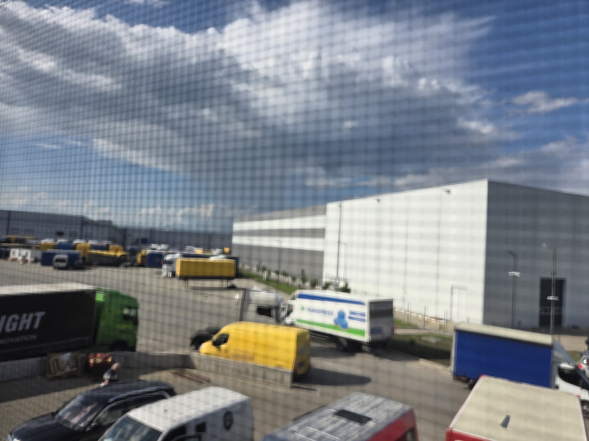 The image, captured through a fine mesh screen, shows a busy industrial or logistics park in Bozhurishte, Bulgaria, under a partly cloudy sky. Large, modern warehouse buildings with white and grey facades form the backdrop. The extensive paved area in the foreground and midground is filled with numerous commercial vehicles.
These vehicles include several delivery trucks and vans: a green-cab semi-truck with a black trailer displaying partial text "IGHT NOVATION" (likely part of "BRIGHT INNOVATION"), various yellow-bodied vans, and a white box truck with the branding "TRANSPRESS" visible in blue and green. A blue box truck and several smaller cars and white vans are also present. One person is vaguely visible standing near a dark car in the lower-left section of the image. The time of day is daytime, with bright natural light creating visible shadows. The weather is partly cloudy, featuring white and grey clouds against a clear blue sky. The scene depicts routine operations at a commercial distribution or shipping hub.