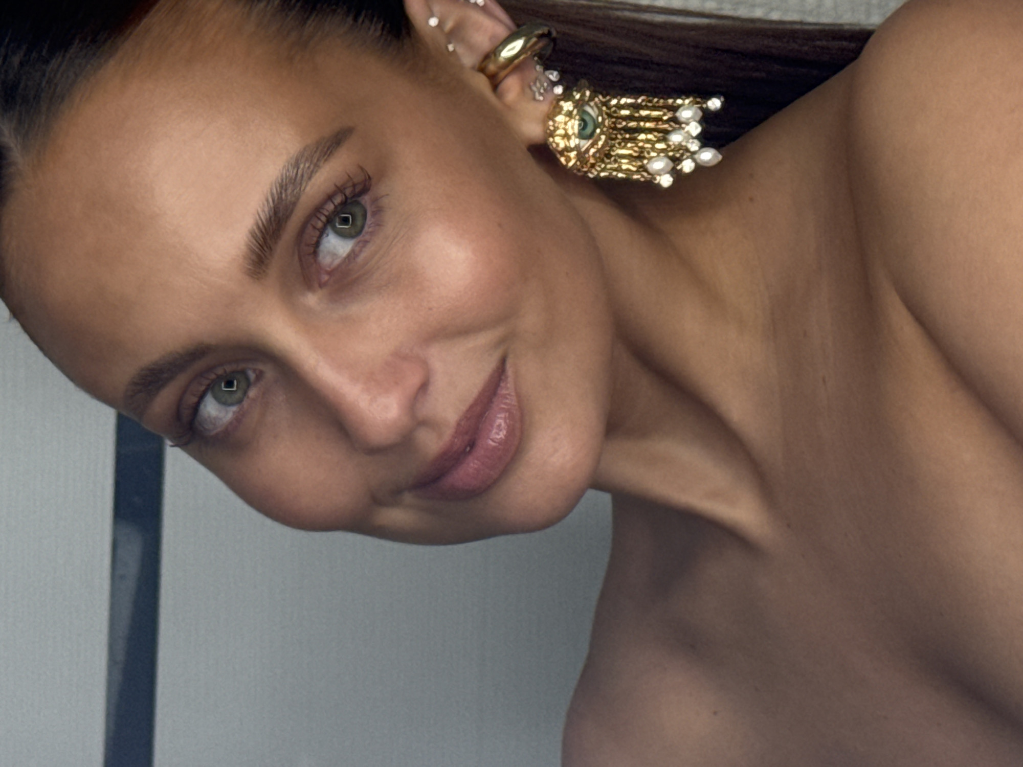 A close-up, angled portrait shows a woman with light olive skin, light green eyes, and light brown hair pulled back. She is wearing a large, gold ear cuff with multiple studs in her ear and a dangling gold earring with a central green stone and pearl fringe. The woman is looking at the camera with a slight smile. Part of a neutral-colored wall is visible in the background. The setting is unknown, but the query mentions the city of Chaikovsky, Russia.