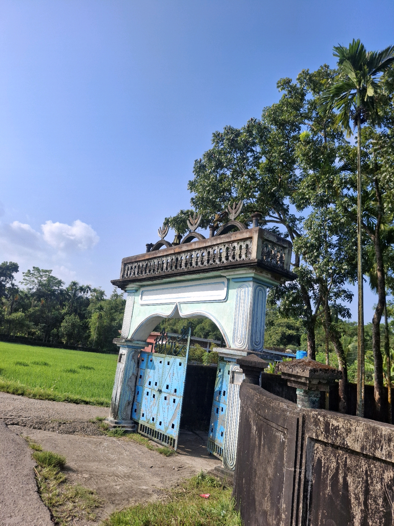 An ornate, weathered gateway stands open, its pastel blue and white arch framing a lush green rice paddy field in Dashghar, Bangladesh. The gateway's upper section features decorative concrete carvings, while the lower section is adorned with fluted columns and a bright blue metal gate with a geometric pattern. A weathered stone wall runs along the right side of the frame, partially obscuring a dense line of trees and palm trees that form the backdrop. The sky is clear blue with a few wispy clouds. The ground in front of the gateway is a mix of dirt and cracked asphalt, with patches of grass growing through. The scene is devoid of people and exhibits a sense of tranquility.