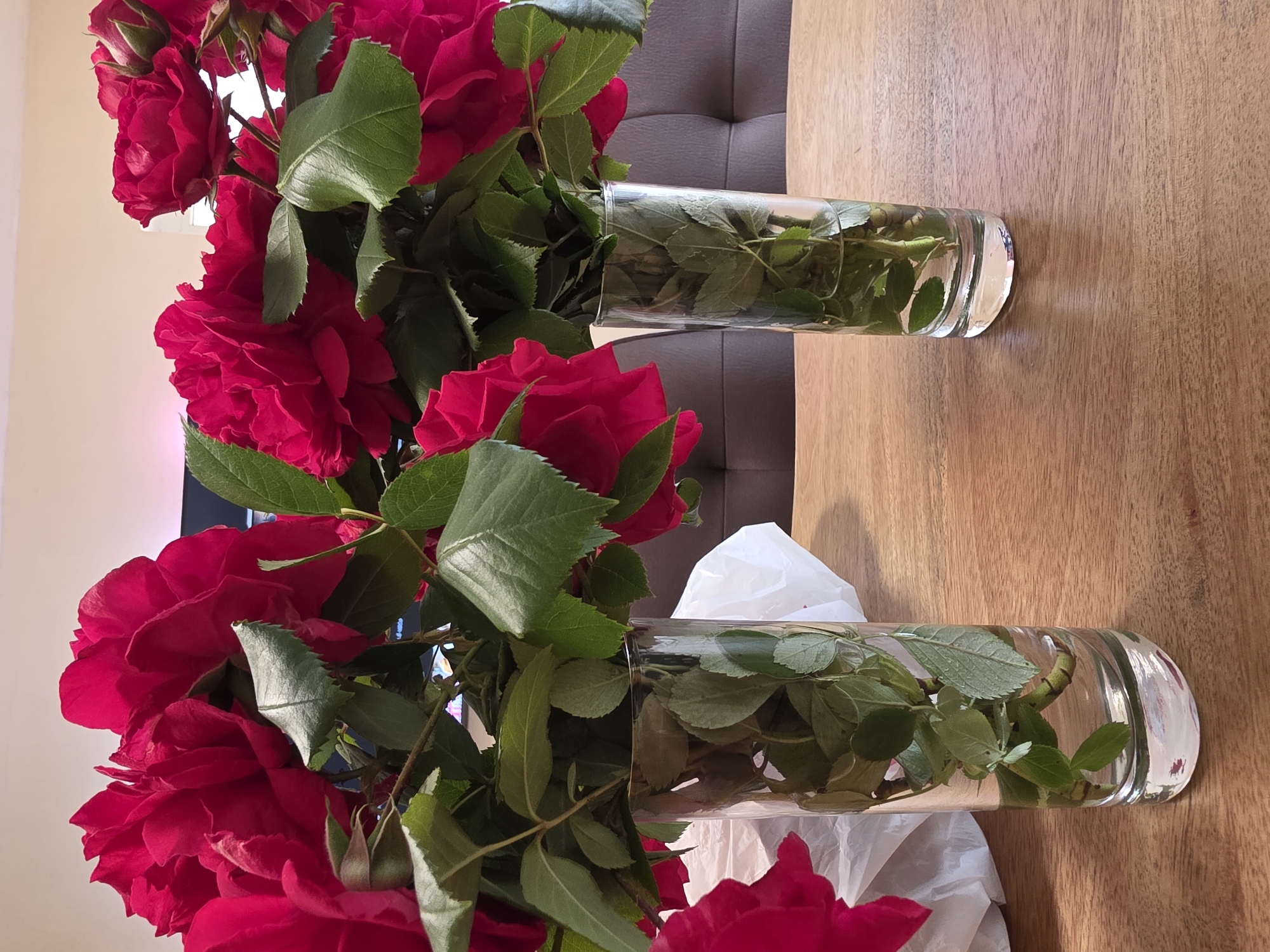 Two clear cylindrical glass vases are arranged on a light brown wooden surface. Each vase contains a bouquet of vibrant red roses with green leaves. The backdrop includes a dark brown cushioned surface behind the vase on the left, and a crumpled white material behind the vase on the right. The scene appears to be indoors. The location in the city of Vinitsa, Bulgaria cannot be determined from this image.