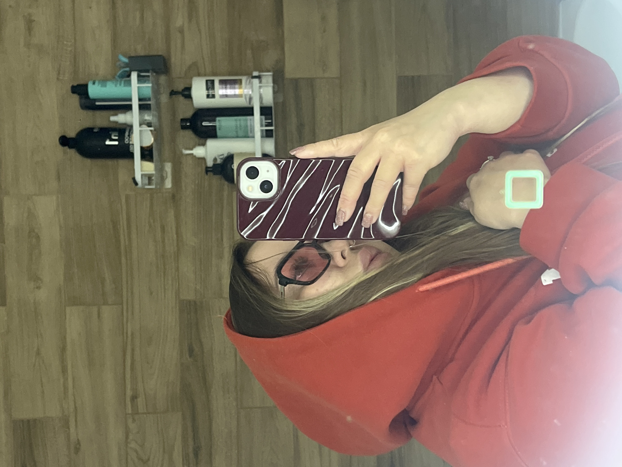The image is a vertical mirror selfie showing a person from the chest up, reflected in what appears to be a mirror. The person is wearing a red hooded sweatshirt, with the hood up, partially obscuring their head. They have long, light brown hair, and are wearing black-framed glasses with reddish-pinkish tinted lenses. Their expression is neutral, partially hidden by the glasses and the angle.
The person is holding a smartphone with their right hand, capturing their reflection. The phone has a dark red or maroon case with white, swirling patterns, and features a dual-camera system on the back. On their left hand, a traditional ring is visible on the ring finger, and a distinct, light green square-shaped ring or device is worn on the index finger.
In the background, behind the person's reflection, there is a clear, multi-tiered shelf attached to a wooden-planked wall or surface. The shelf holds several bottles, likely hair or body care products, in various colors including light blue, black, and white. Text on one black bottle clearly reads "SHAMPOO GEL." Other bottles have partially visible labels, but the text is not fully legible.
The setting appears to be indoors, possibly a bathroom or dressing area, indicated by the arrangement of the personal care products and the wooden paneling. The lighting is bright, creating a slight glare on the mirror, particularly on the right side of the image. The overall activity is a casual self-portrait taken in a personal space. This scene takes place in Krasnodar, Russia.