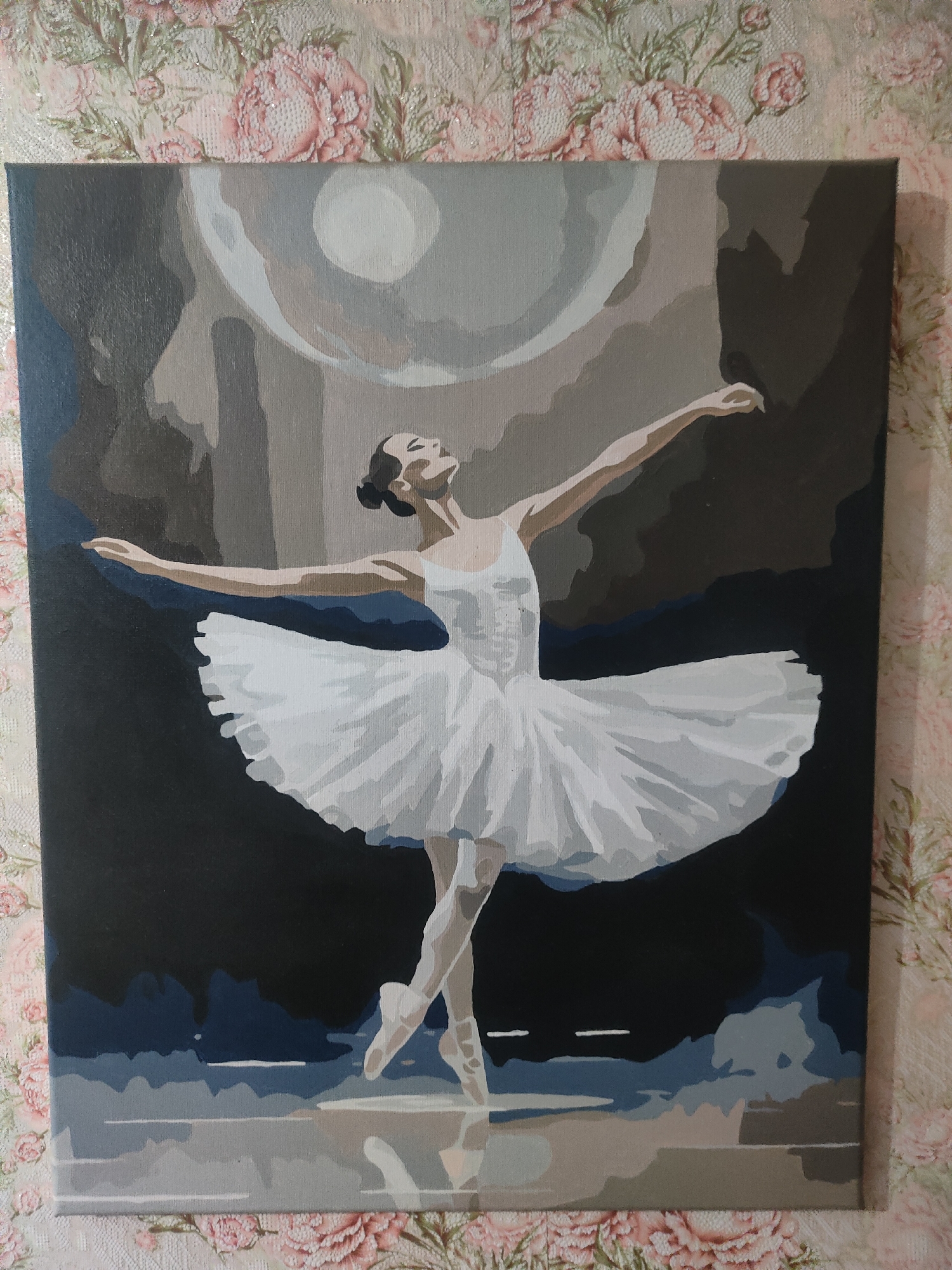 A painting depicts a ballet dancer in a white tutu and bodice, poised en pointe with arms extended. The dancer's head is tilted back, and her eyes are closed. The background is abstract, with shades of gray, blue, and brown, suggesting a stage or a moonlit sky. The painting is displayed against a floral-patterned wallpaper. The location is identified as Ternovka, Moldova.