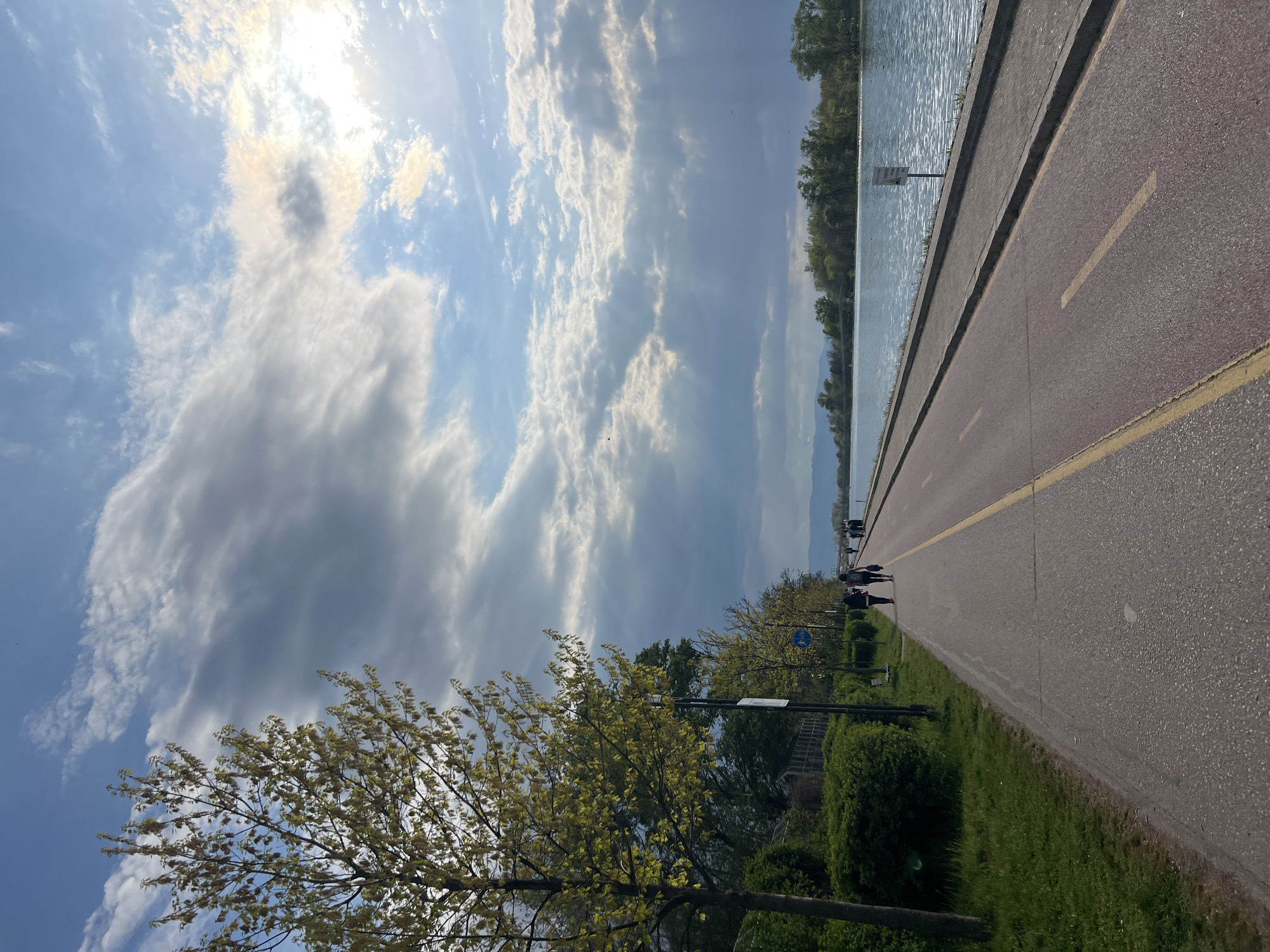 This media file presents a scenic waterside pathway, likely in Plovdiv, Bulgaria, captured during what appears to be a bright, partly cloudy day. The original image is rotated, but when viewed horizontally, it depicts a long, paved path stretching into the distance, with a body of water to its right and green foliage to its left.
**Subjects:**
The primary subjects include a wide, asphalt pathway with a prominent yellow dividing line, suggesting separate lanes for pedestrians and cyclists. To the right of the path, a calm body of water extends towards a distant tree-lined bank and mountains. On the left, lush green grass, bushes, and trees with fresh, light-green spring leaves line the path. Several individuals are visible walking leisurely along the path in the mid-ground, enjoying the setting. The sky occupies a significant portion of the upper frame, filled with a mix of bright white and darker gray clouds, with the sun brightly shining through, creating visible sunbeams.
**Activity or Event:**
The scene captures people engaged in a recreational activity, such as walking or jogging, along a scenic waterfront promenade. The overall atmosphere suggests a leisurely outdoor experience.
**Notable Details:**
The time of day appears to be late morning or afternoon, characterized by the bright sunlight reflecting off the water and illuminating the clouds. The weather is pleasant, with a clear blue sky interspersed with scattered clouds. A blue circular sign with a white bicycle symbol is clearly visible on the left side of the path, confirming it is a designated cycling route. A small, unreadable white sign is also present near the water's edge further down the path. The vibrant, light green leaves on the trees indicate springtime. The distant mountains provide a picturesque backdrop to the tranquil water.
**Visible Text:**
The only clearly discernible text is the white bicycle symbol on the blue circular sign.