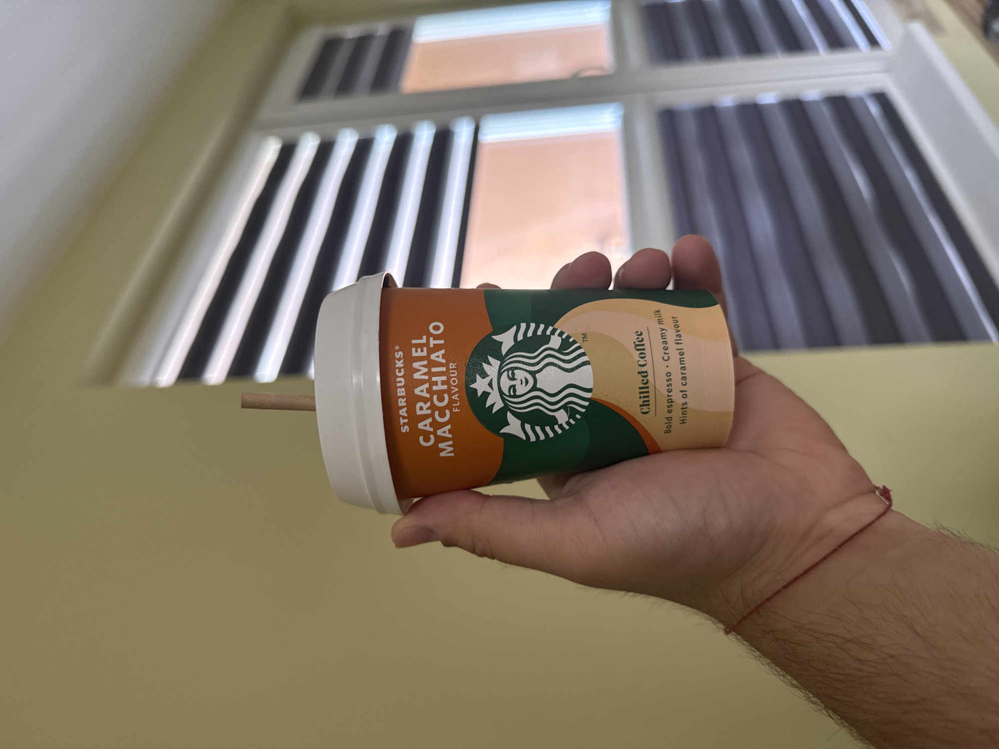 A person's hand is holding a Starbucks Caramel Macchiato chilled coffee cup. The cup is orange and green with the Starbucks logo and text. A small brown straw is visible sticking out of the top. The background shows a wall and a window with horizontal blinds. The scene appears to be indoors.