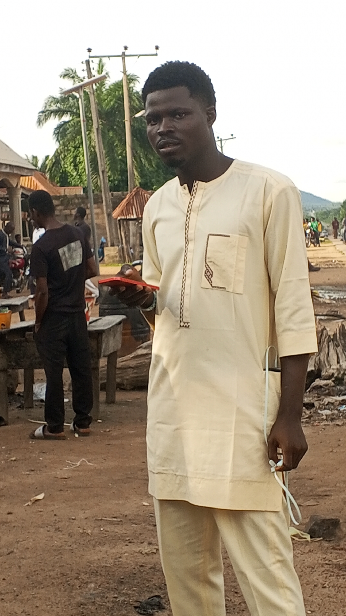 A full-shot image shows a young Black man wearing a light yellow, traditional Nigerian outfit, including a long-sleeved tunic with a patterned neckline and pocket, and matching pants. He is standing in a dirt-covered outdoor area with a blurred background that includes buildings, trees, and utility poles. In his right hand, he holds a red object, possibly a phone, and in his left hand, he is holding a white earbud. His gaze is directed towards the viewer. To his left, another man in a black t-shirt and pants is visible, also looking away from the camera. The environment appears to be a bustling marketplace or street in a Nigerian city, likely Nyita, given the context of the request.