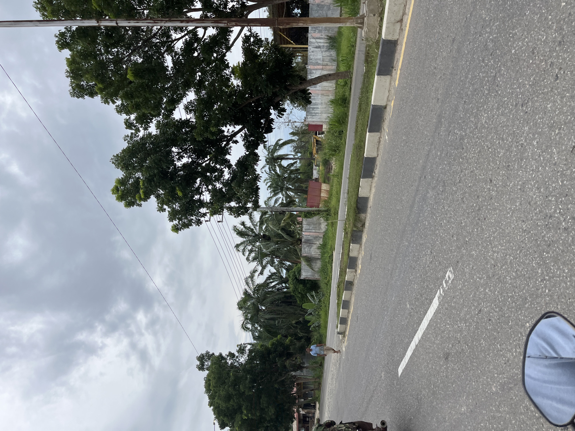 A man walks on the side of a road in Tanahterban, Indonesia. He is wearing a light blue short-sleeved shirt and light-colored shorts. The road is paved and has a white line marking the lane. The man is walking away from the viewer, towards the right side of the frame. The area is lined with trees, some of which are palm trees. In the background, there are buildings and construction equipment, including a yellow excavator. The sky is overcast with gray clouds. The image appears to have been taken from a moving vehicle, possibly a motorcycle, as a portion of a side mirror is visible in the lower left corner of the frame.