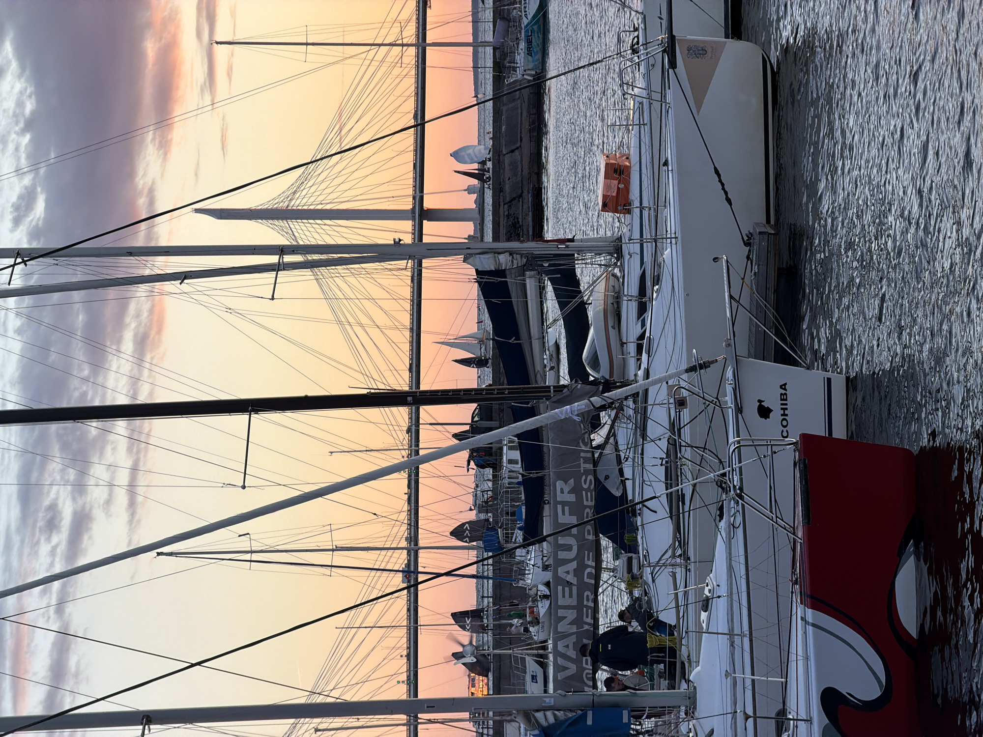 The image depicts a marina in Saint Petersburg, Russia, likely near the Western High-Speed Diameter (WHSD) bridge, as indicated by the distinctive cable-stayed bridge visible in the background. The scene is set at sunset, with a warm, orange-pink hue illuminating the sky and reflecting on the water. Numerous sailboats are moored in the foreground and midground.
In the midground, several sailboats are clearly visible. One boat has a prominent banner with the text "VANEAU.FR" and "JOELIER DE PRESTIGE". On this boat, two individuals are standing on deck, seemingly engaged in conversation. Another sailboat to the left has a red and white hull with stylized wave-like graphics. Further back, other sailboats with sails partially furled or unfurled are visible.
In the foreground, the sterns of several sailboats are prominent. One white sailboat has the name "COHIBA" written on its stern, along with a silhouette of a head. Another white sailboat to its right has a crest-like emblem. The water is calm with ripples reflecting the colors of the sky.
The overall atmosphere is serene, capturing a moment of repose for the sailboats and their occupants as the day concludes. The presence of the WHSD bridge anchors the location to Saint Petersburg.