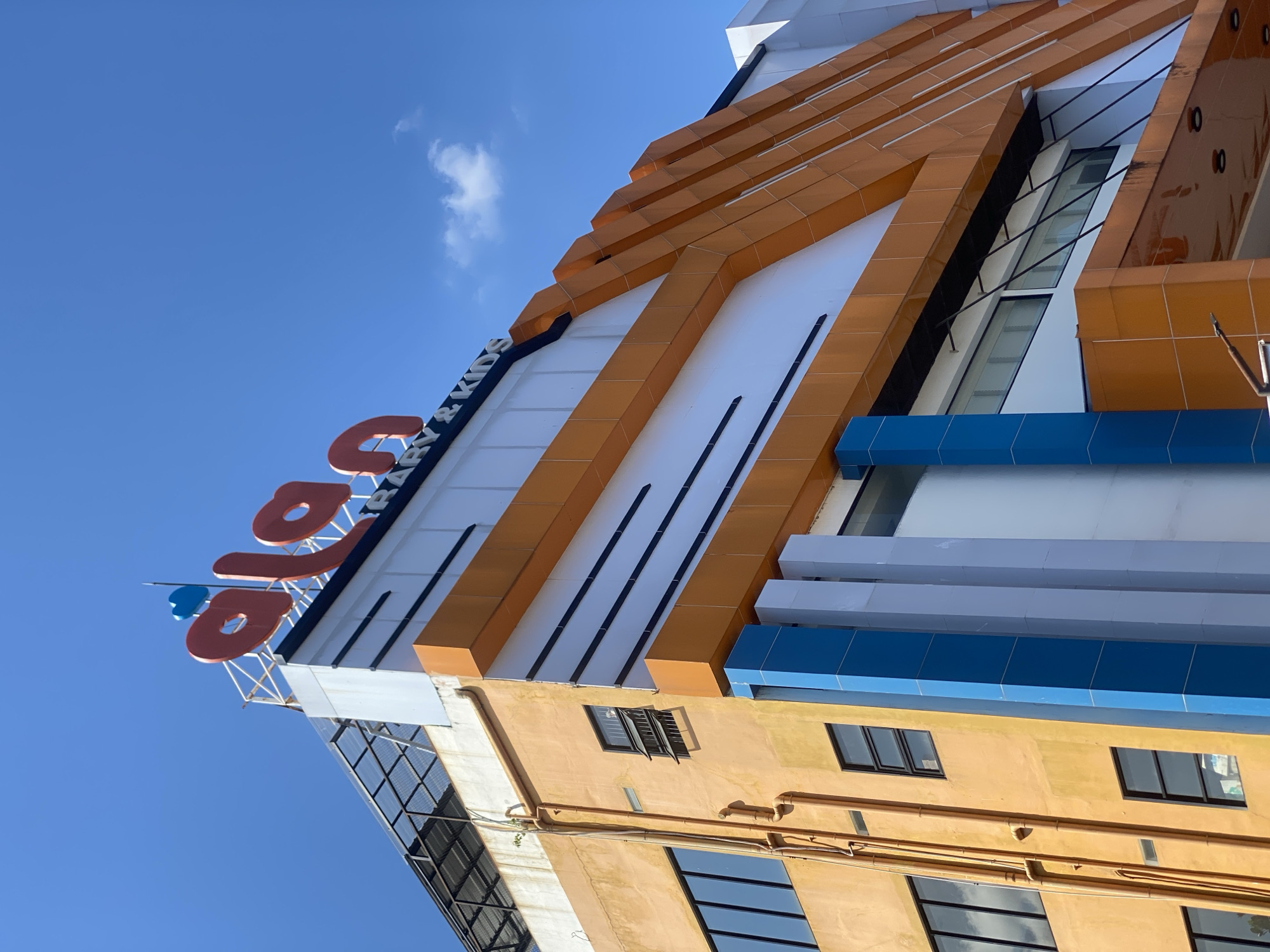 The image shows the exterior of a building in Sungai Paring, Indonesia. The building has an orange facade with blue and white accents. A sign at the top of the building reads "alan" in large red letters, with "PRIMARY & KIDS" written below in smaller letters. The sky is blue with a few scattered clouds. The building appears to be a commercial establishment, possibly a school or a children's center. The image is taken from a low angle, looking up at the building. There are no people visible in the image.