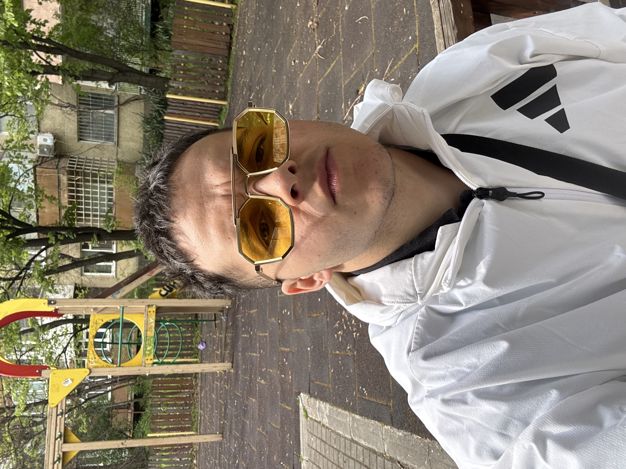 A person is depicted in an outdoor setting, likely a park or residential courtyard, with the image rotated 90 degrees counter-clockwise. The main subject is an individual, seen from the chest up, wearing large, gold-framed octagonal sunglasses with yellow-tinted lenses. They are dressed in a white Adidas jacket, featuring the prominent black three-stripe logo on the shoulder/upper arm area, and a black strap is visible across their chest.
The background reveals a playground to the left, equipped with yellow and red plastic/metal structures and green metal rings, suggesting a family-friendly environment. Behind the playground are residential buildings, characterized by windows, some with security grates, and an visible air conditioning unit. Mature trees with green foliage are scattered throughout the scene, indicating spring or summer. The ground is paved with rectangular stone or brick pavers.
The scene takes place during the daytime, under bright sunlight, as evidenced by the clear illumination and shadows. The person appears to be posing for a photograph or taking a selfie. A small yellow sticker with "ELVE" written on it can be faintly seen on one of the yellow playground structures. The combination of residential buildings, a playground, and urban paving aligns with the specified location context of Sofia, Bulgaria.