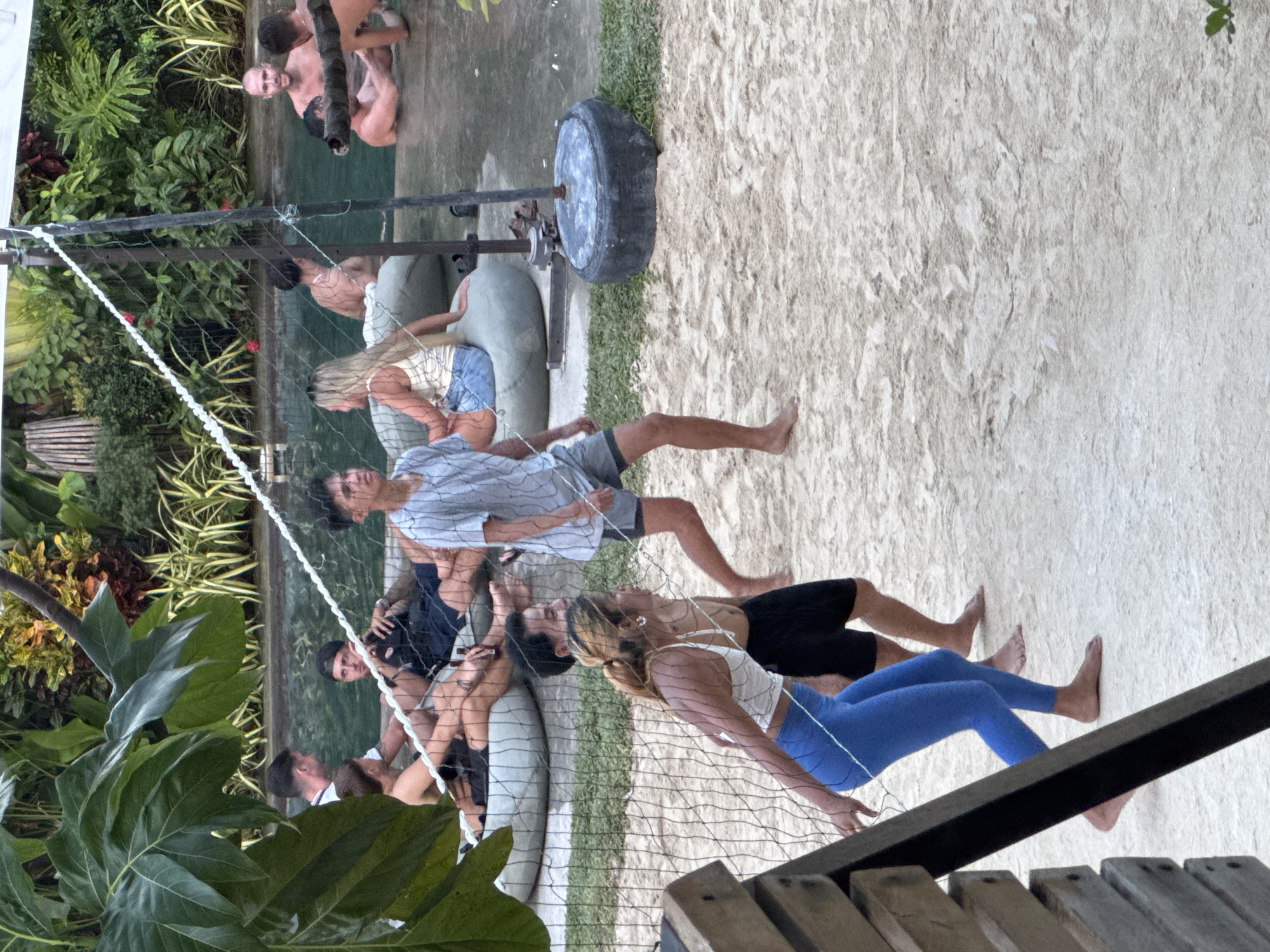 A horizontal medium shot depicts a group of individuals lounging on cushioned seating behind a volleyball net, situated on a sandy area that possibly serves as a makeshift beach. The scene is in General Luna, Philippines.
In the foreground, the bottom of a wooden structure with visible planks leads into the scene from the lower right corner. A woman with light hair is closest to the viewer, wearing a white top and blue leggings. Adjacent to her is a man in black shorts. Further back are more individuals seated on cushioned surfaces. A person in a grey shirt and grey shorts stands barefoot in the sandy area. Behind the people in seats is a volleyball net supported by metal posts. A tire rests on a sandy area near the posts. Beyond the volleyball setup, lush green plants and trees surround the perimeter of the space, and shirtless individuals are on the edge. The ground is covered in sand.