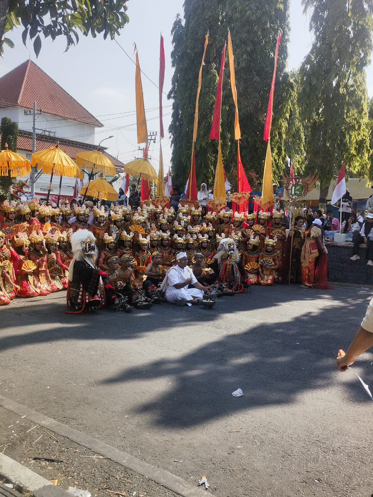 This is a medium-shot, eye-level, outdoor, daytime photograph of a cultural performance taking place in Blora, Indonesia. The foreground shows a paved street with a concrete curb and scattered debris. A large group of performers, dressed in traditional Balinese attire, are arranged in rows, some seated and others kneeling. The performers are adorned with elaborate headdresses, colorful costumes, and some are holding fans. In the front left, a performer in a striking mask and ornate costume stands out. To the right of the masked performer, another individual in white attire is seated on the ground. Behind the main group of performers, several large yellow umbrellas are visible, along with tall trees and buildings. Red and yellow flags are also prominently displayed, some of which are attached to poles and appear to be part of the procession or performance. The overall scene suggests a festive cultural event.