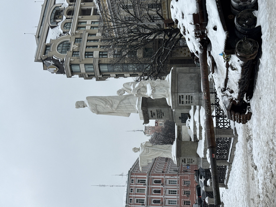 An overcast winter scene in Kyiv, Ukraine, depicts the Monument to Magdeburg Rights on Volodymyrska Hill, contrasting with a destroyed tank. The monument features a central, tall white statue of Volodymyr the Great on a grey pedestal, flanked by two smaller white statues: one of Saints Cyril and Methodius with an outstretched arm, and another of Nestor the Chronicler seated with a book. All monument figures and their pedestals are lightly dusted with snow. A dark, ornate metal fence encircles the monument's base.
In the foreground to the right, a heavily damaged and rusty tank wreck rests in thick snow. Its main gun barrel extends horizontally to the left, displaying white spray-painted Cyrillic letters "ХОРУ." A small blue and yellow Ukrainian flag is affixed to the tank's side.
In the background, a modern, multi-story building with a curved facade, a snow-covered roof, and a prominent golden "1" logo stands to the right of the monument. To the left, an older, reddish-pink multi-story building with white architectural details is visible. Bare deciduous trees are interspersed within the urban environment. The ground is entirely covered in a layer of uneven snow. No individuals are present.