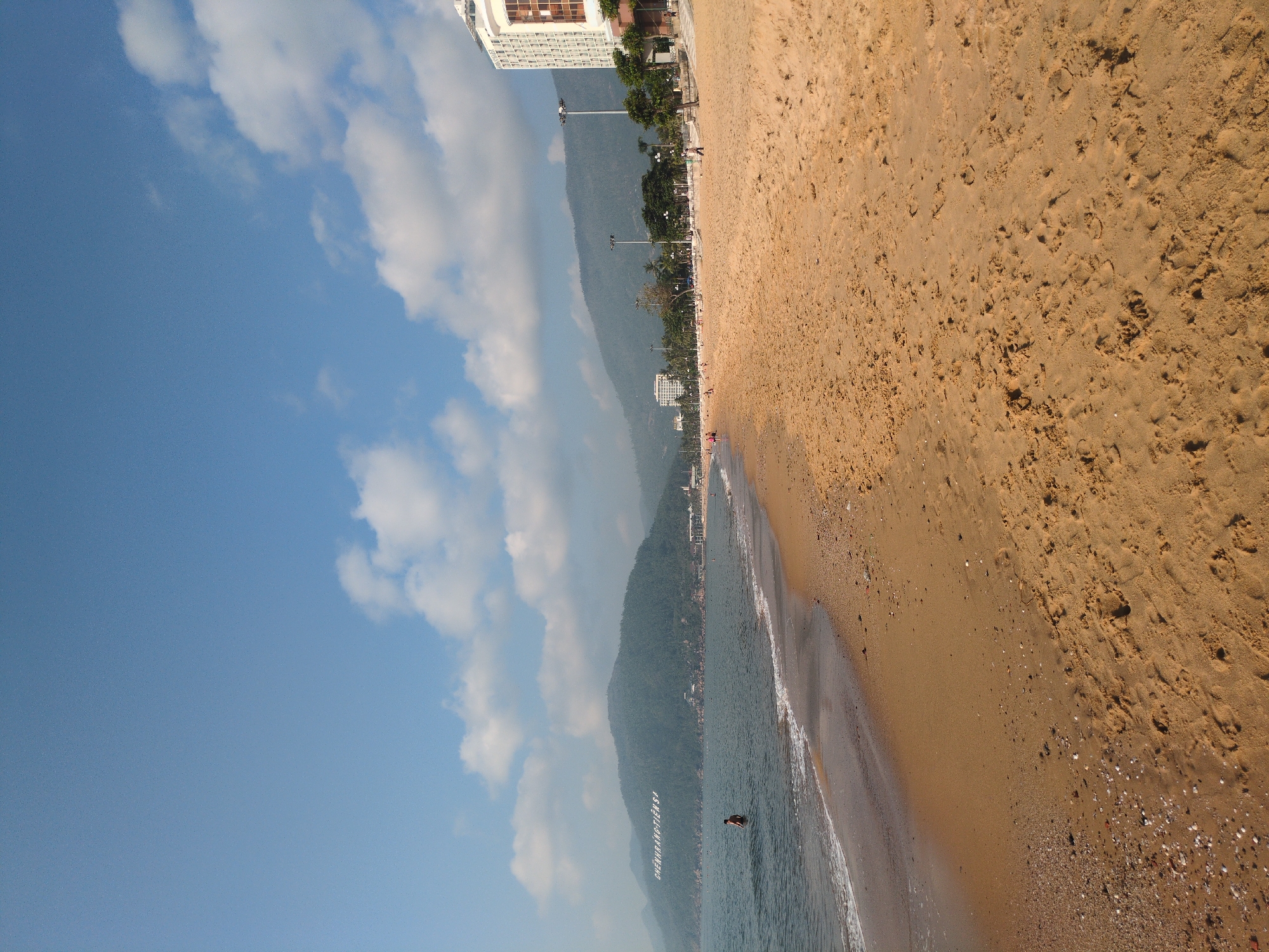 The image captures a beach scene in South Quy Nhon Ward, Vietnam. To the left, a blue sky is partially filled with white clouds. A series of mountain peaks extends along the coastline. Adjacent to the mountains are buildings, including a large hotel, lining a street with palm trees. The ocean water meets the sandy beach in the foreground. A single person is wading in the water. The sand shows signs of footprints and varying textures.