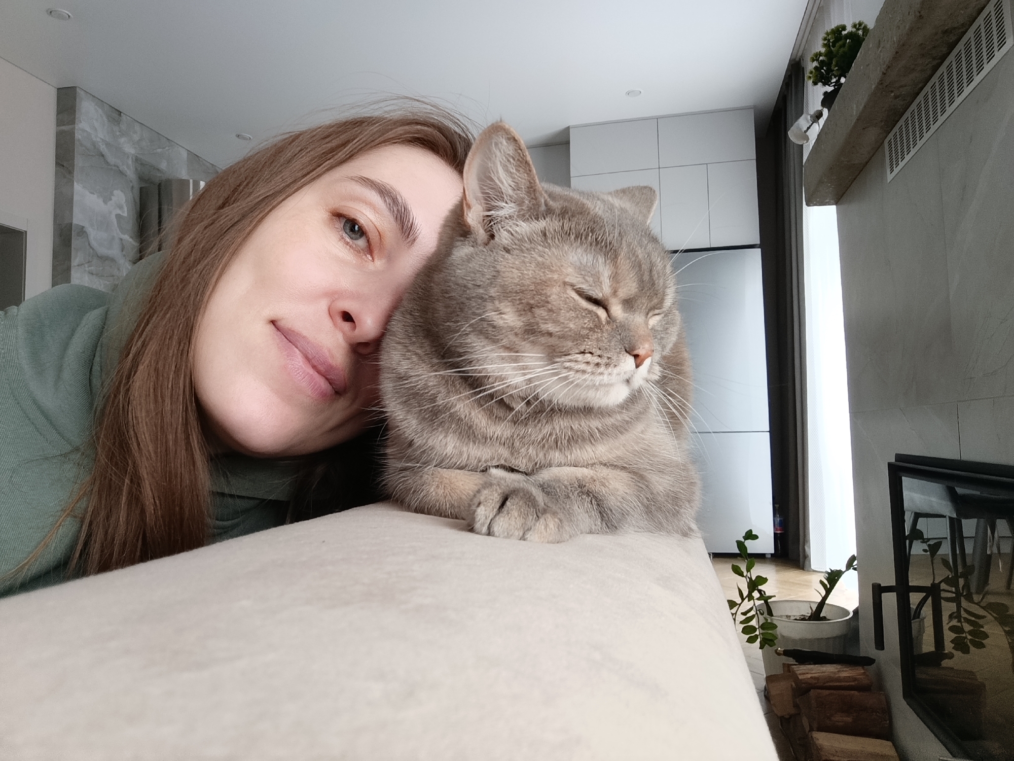 A heartwarming close-up selfie captures a young woman and her grey tabby cat sharing a peaceful moment in a modern interior, likely in Chaikovsky, Russia. The woman, with light brown hair and blue eyes, leans her head gently against her feline companion, displaying a soft, contented smile. She is wearing a sage green hooded top. The grey tabby cat, possibly a British Shorthair, rests its head and paws on a light-colored sofa or cushion, with its eyes closed in a state of serene relaxation or sleep. Its striped fur and prominent white whiskers are clearly visible.
The background reveals a contemporary living space, bathed in natural daylight, suggesting it is daytime. To the left, sleek white kitchen cabinetry or large appliances are visible, alongside wall panels with a subtle grey marble pattern and recessed ceiling lights. On the right, elements of the room include a dark-framed mirror or fireplace, below which a small stack of firewood logs is neatly arranged next to a potted green plant. Above the fireplace, a stone-like shelf holds another small potted plant and an inverted glass light fixture, with a white ventilation grille integrated into the wall nearby. Bright light streams in from a tall window on the far right. The overall ambiance is one of calm affection and domestic comfort. No visible text is present in the image.