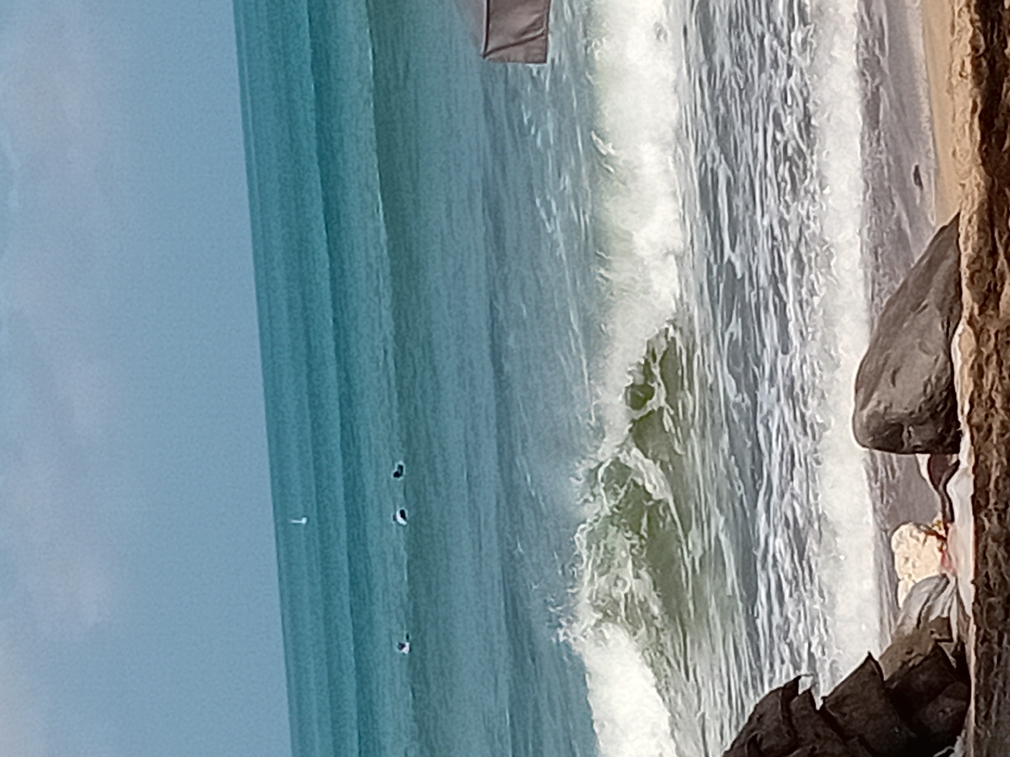 The image captures a view of the ocean from the shore in Kuta, Indonesia. In the distance, a small sailboat is visible on the calm blue water. Closer to the shore, three surfers are positioned on their boards, appearing to be waiting for waves. A large wave is breaking in the mid-ground, with white foam and spray. The foreground features a rocky shoreline with a large dark boulder on the left and a lighter-colored rock and some debris on the sand to the right. The sky is overcast with a light blue-grey hue.