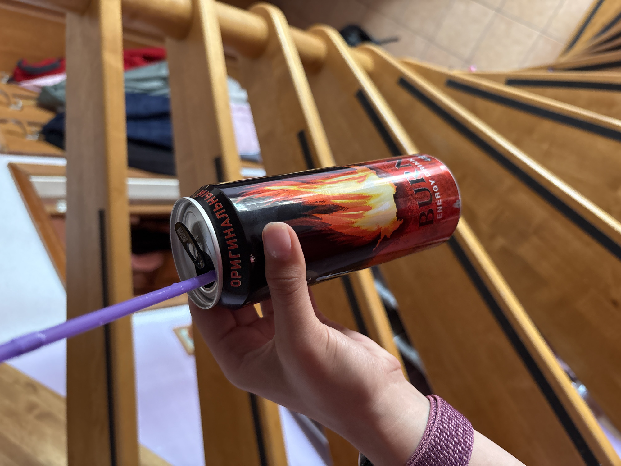 A hand holds a can of BURN energy drink with a purple straw inserted. The can is black and red, with a flame design. The hand has a purple woven bracelet. The background shows a light wooden spiral staircase. The text "ОРИГИНАЛЬНЫЙ" is visible on the can. The location might be Прикамский, Russia.