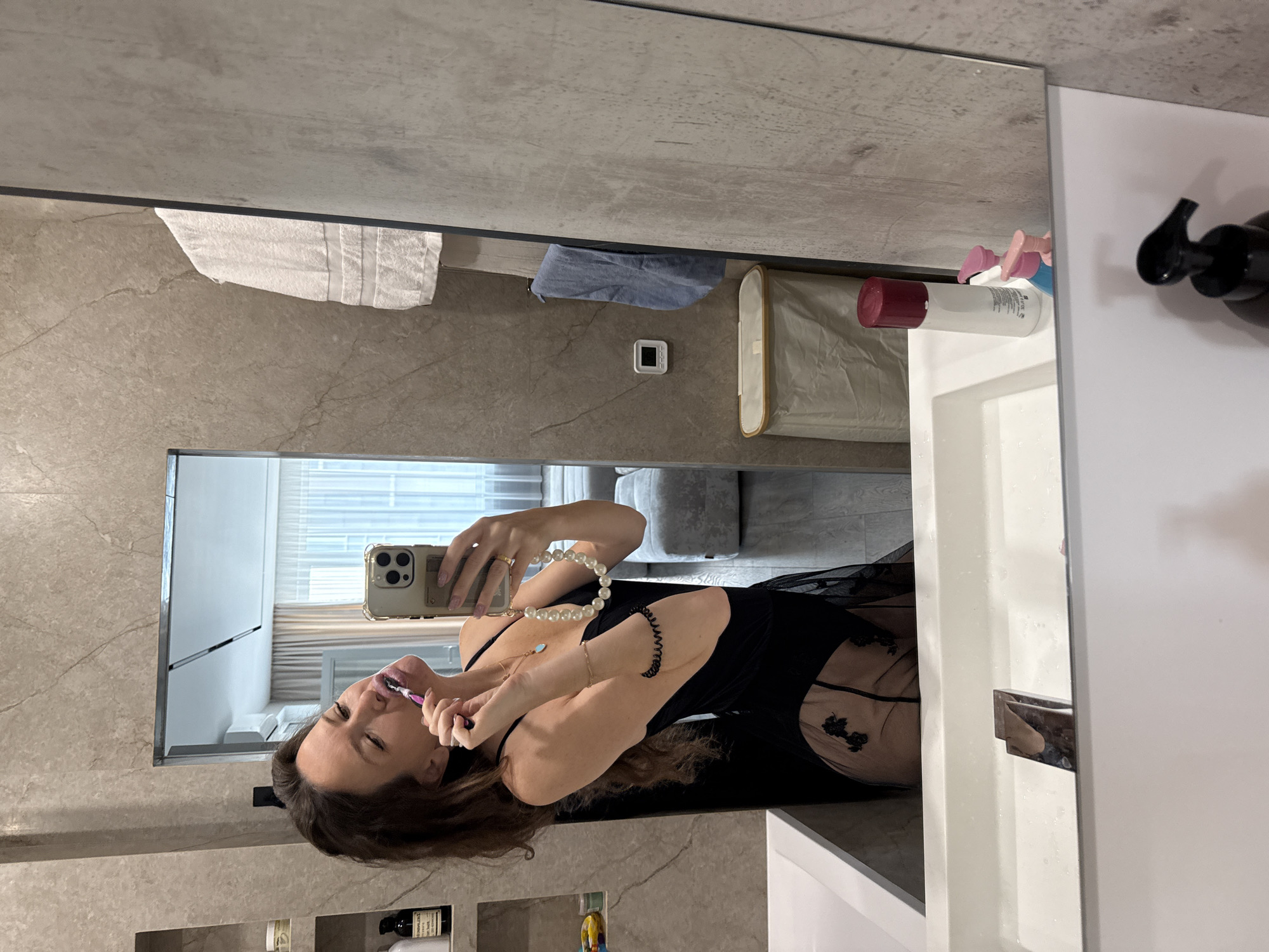 A person is standing in front of a mirror in a bathroom and taking a picture of themselves while brushing their teeth. The bathroom has marble-like walls and a white sink. Various toiletries are visible next to the sink. In the reflection of the mirror, a living area with a couch is seen. The exact location in Khimki, Russia cannot be determined from the image.
