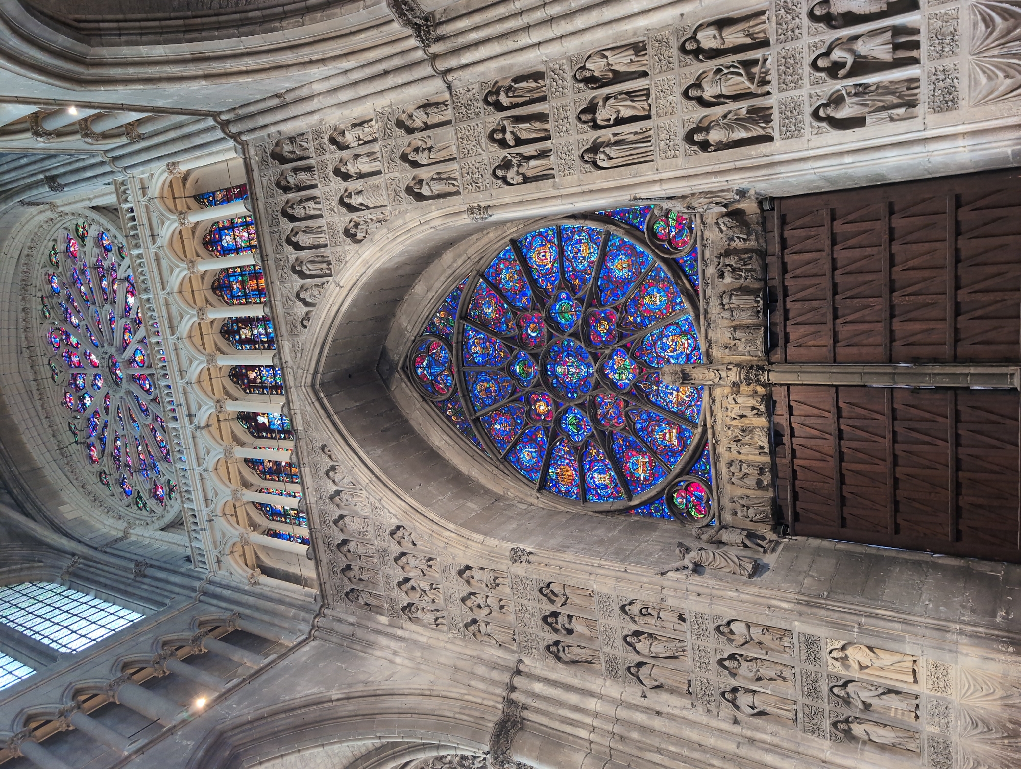 The image shows a view of a grand cathedral interior, with a large rose window dominating the center of the frame. The rose window is made of stained glass and features intricate patterns in shades of blue, red, and purple. Below the rose window are massive wooden doors, ornately carved and set within a stone archway. The walls of the cathedral are adorned with numerous stone sculptures of figures, arranged in rows within arched niches. The architecture is Gothic, characterized by pointed arches, ribbed vaults, and soaring ceilings, with sunlight filtering through the stained glass, casting colorful patterns on the stone. This is the interior of the Cathédrale Notre-Dame de Reims, located in Reims, France, not Urvillers, France.