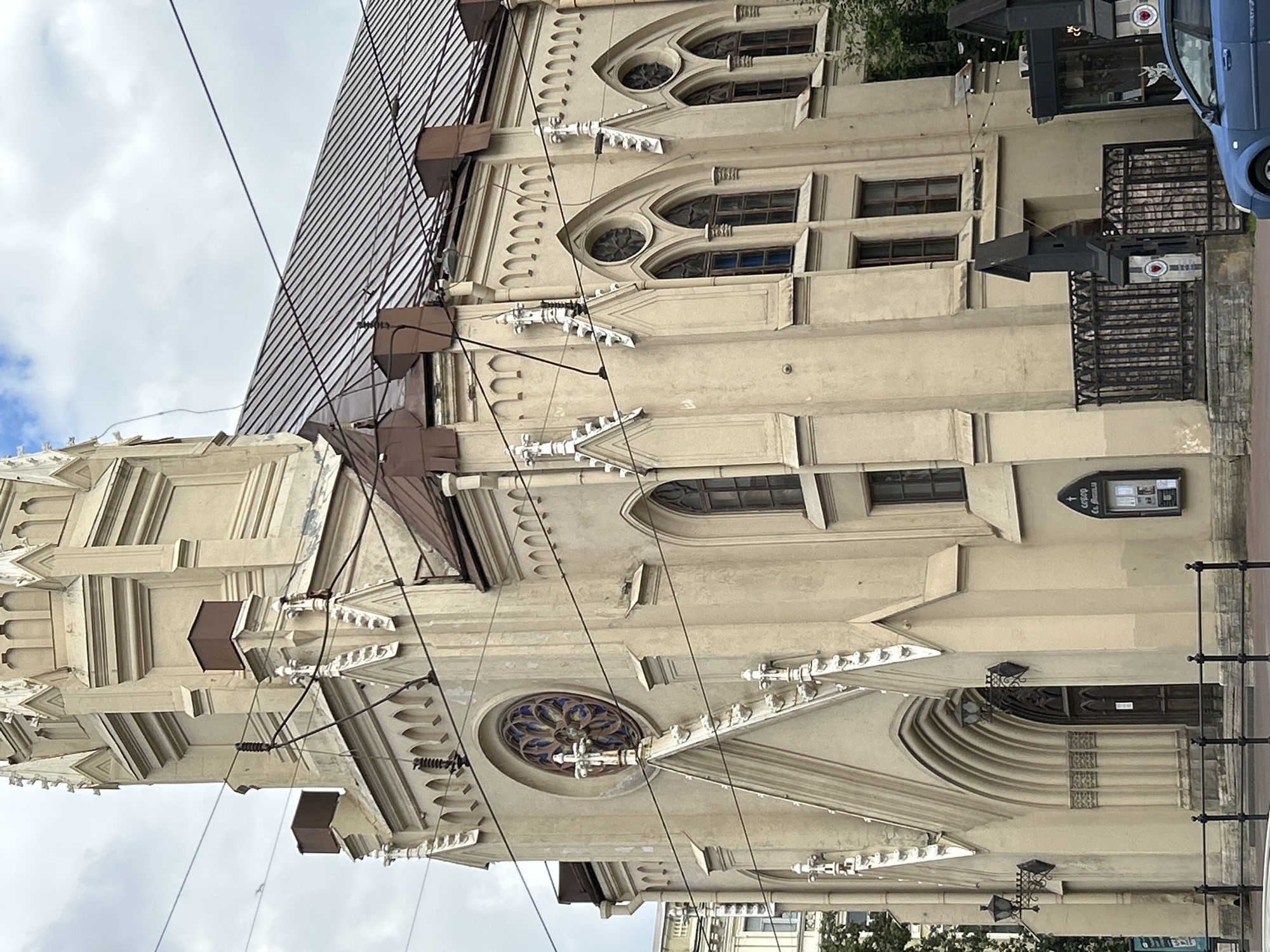 The image shows a tan-colored Neo-Gothic building in Saint Petersburg, Russia, likely a church or a similar historical structure, with intricate architectural details. The building features pointed arch windows, decorative spires, and a large rose window above the main entrance. The roof is made of dark, ribbed material. Utility wires crisscross the sky in front of the building, suggesting an urban environment with overhead electrical infrastructure. A dark wooden door is visible beneath the rose window, accessed through a large pointed archway. To the right of the entrance, a dark metal fence encloses an area with a stone base and decorative pillars. A blue car is partially visible in the foreground on the right side of the frame. The sky is partly cloudy, indicating an overcast day.