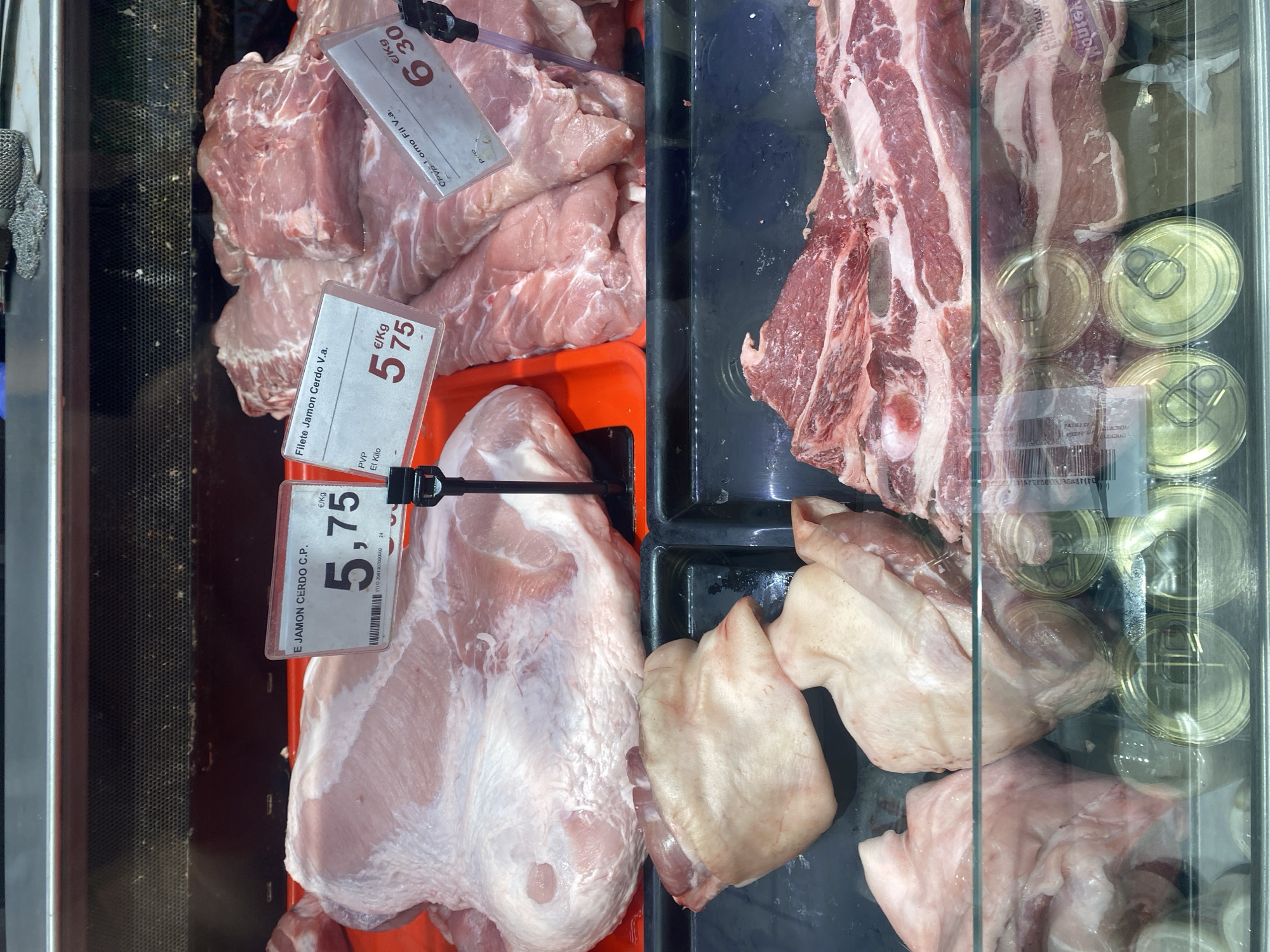 The image shows a close-up of a display case filled with various cuts of raw pork in a butcher shop. The lighting is bright, illuminating the fresh meat. In the foreground, on the left, there are several pieces of pork, some with skin and fat. To the right of these, a large cut of pork is visible, with a label indicating "E. JAMON CERDO C.P." and a price of "5,75 €/Kg". Further to the right, another cut of pork, identified as "Filete Jamon Cerdo V.a.", is priced at "6,30 €/Kg". In the background, more raw pork cuts are displayed. Below the meat, in the lower part of the image, metallic cans are visible through a glass partition. The overall environment appears to be a commercial setting for selling meat products. The location is specified as Bel Air, Spain.