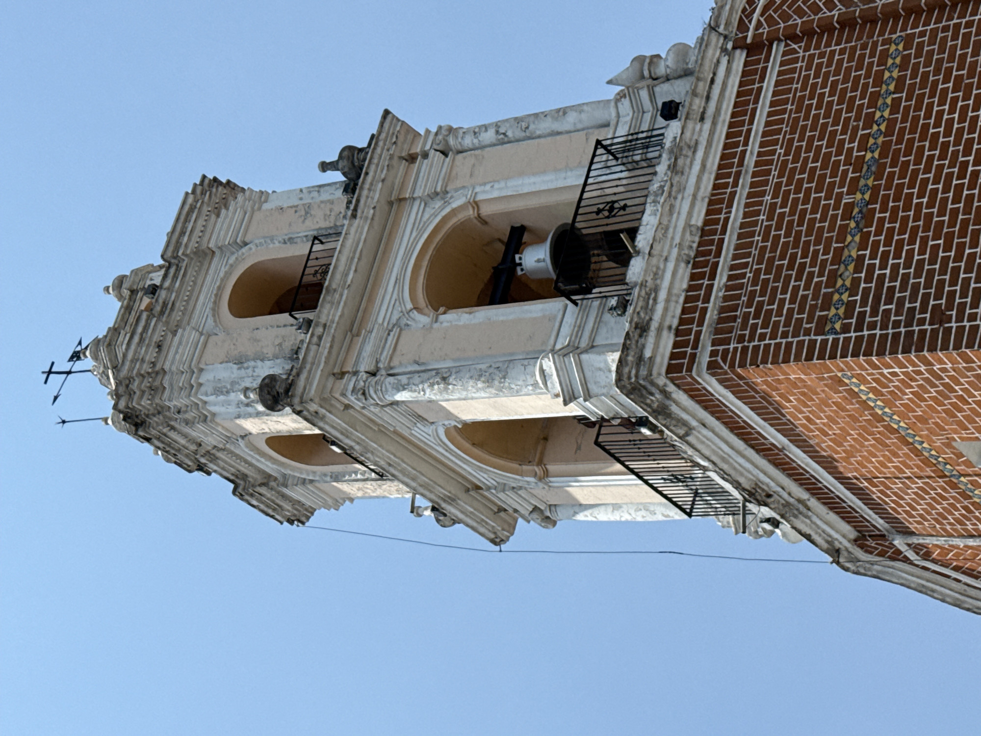 An ornate bell tower or church facade is captured from a low-angle perspective, viewed against a clear, bright blue sky. The image is oriented sideways, but depicts a vertical structure. The tower is constructed in multiple tiers, featuring arched openings, at least one of which reveals a large metal bell, primarily white with black supports. Decorative black metal railings are installed in front of these openings.
The architectural style includes detailed cream-colored or light-painted stone and stucco elements such as cornices, moldings, and decorative carvings that show signs of weathering. Protruding from the corners of the upper sections are sculptural elements, possibly gargoyles or decorative finials. At the very top, a slender weather vane or lightning rod is visible against the sky.
A prominent feature is the right side of the structure, which is a wall of red brick laid in a stretcher bond pattern with white mortar. Running vertically along this brick wall is a distinctive decorative band composed of small, patterned tiles in yellow, blue, and white, suggesting local artisanal work like Talavera. The bright, cloudless sky indicates a sunny day. There are no people or discernible activities, and no visible text. The combination of brickwork, patterned tiles, and architectural style aligns with structures found in Puebla, Mexico.