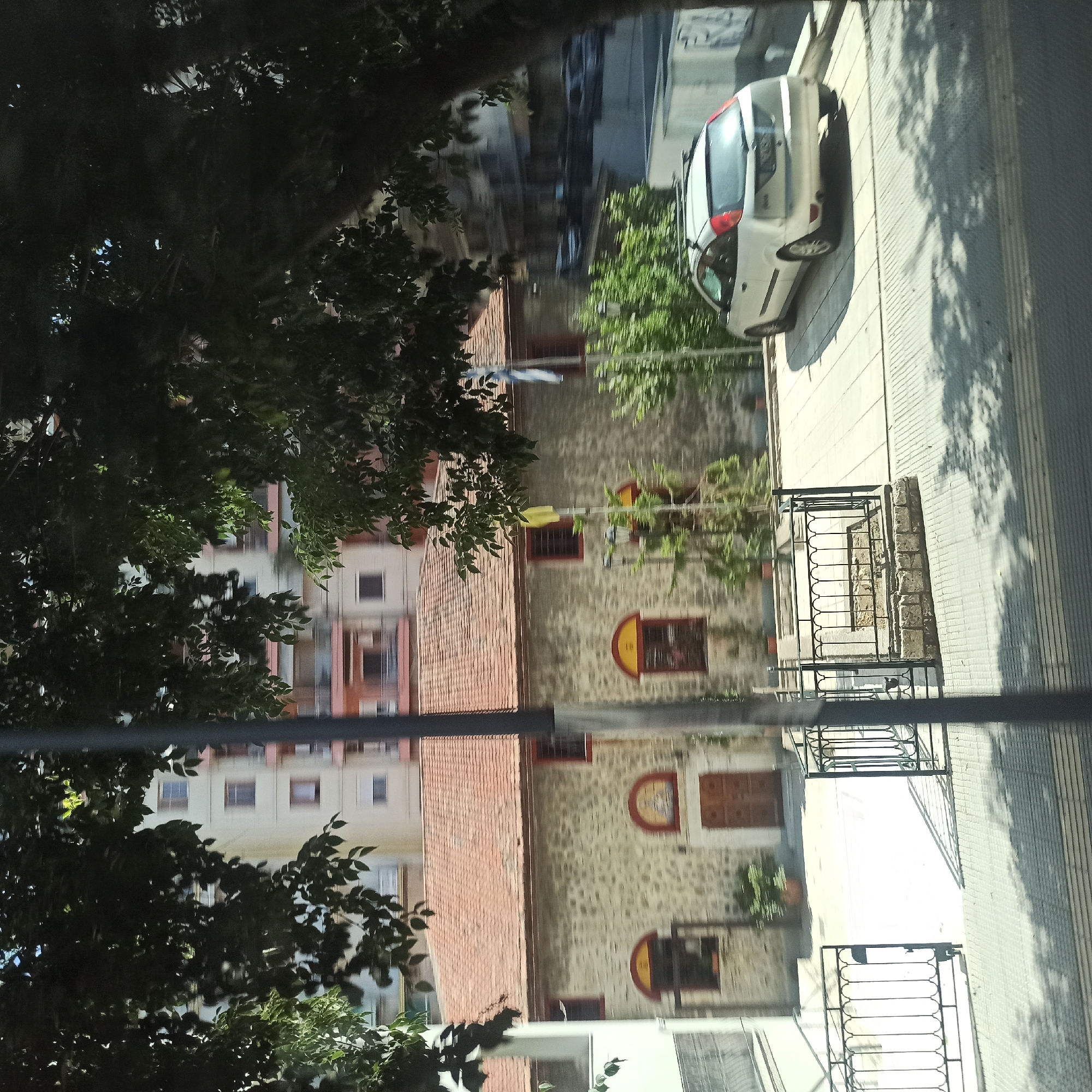 This is a photo taken from a moving vehicle, likely a bus, in Thessaloniki, Greece. The image captures a glimpse of a stone building with a red-tiled roof, possibly a church, with arched windows and wooden doors. In front of the building, a white car is parked. The scene is partially obscured by the leaves and branches of a large tree in the foreground. The background shows a multi-story residential building. The overall impression is of a sunny day in a Mediterranean city.