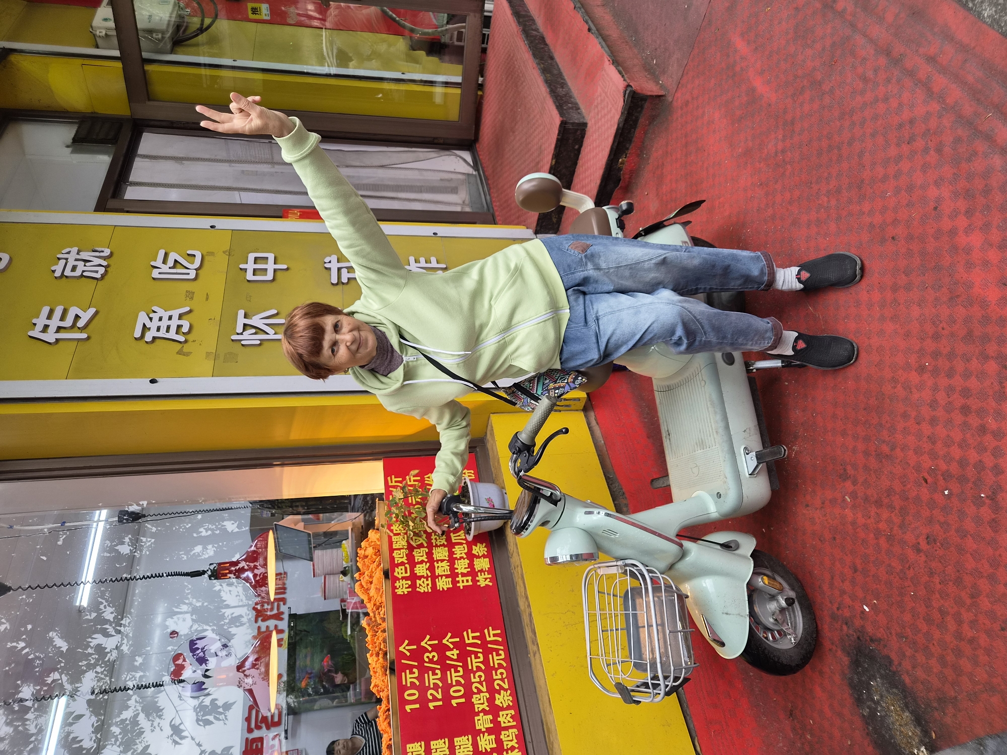 A smiling elderly woman with short, reddish-brown hair is sitting on a light green electric scooter parked on a bright red textured surface. She is wearing a light green hooded jacket and blue denim jeans. Her left arm is raised, and she is making a peace sign with her fingers. A patterned bag is slung over her right shoulder. The scooter has a front basket and a brown seat. Behind her, a yellow building with Chinese characters is visible. A red sign with Chinese text and prices is displayed on the building, advertising food items such as chicken. A glass window shows a display of fried food and what appears to be a goldfish tank. Red steps lead up to a doorway on the right side of the building. The location is identified as Mishan, China.