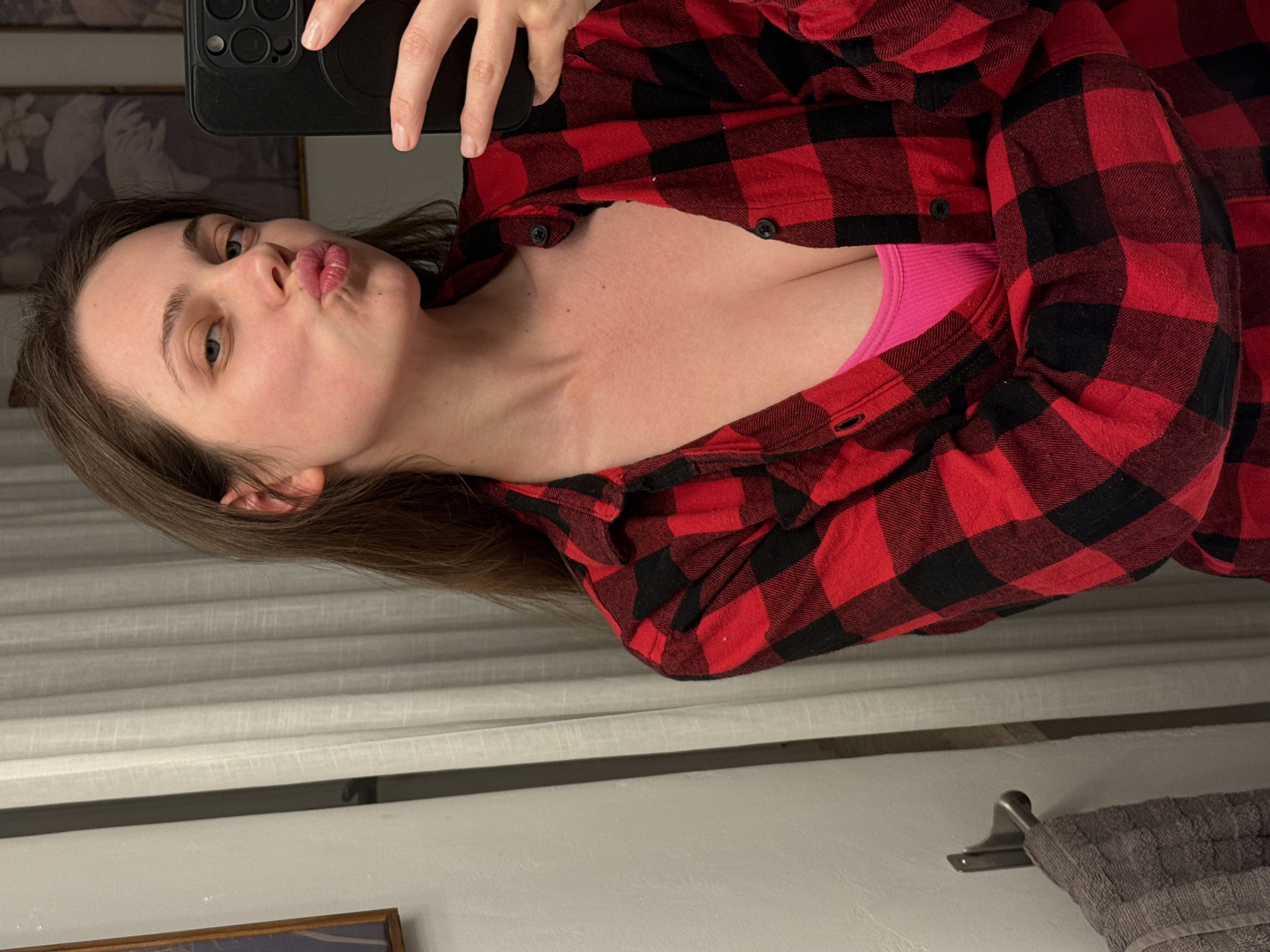 A woman with brown hair is taking a selfie in a bathroom mirror. She is wearing a red and black plaid shirt over a pink top. Her lips are pursed in a kissing gesture. The bathroom features light-colored walls, a towel rack with a gray towel, and blinds on a window. Part of a framed picture is visible in the bottom left corner. The scene appears to be indoors, and there is no indication of it being in Perugia, Italy.