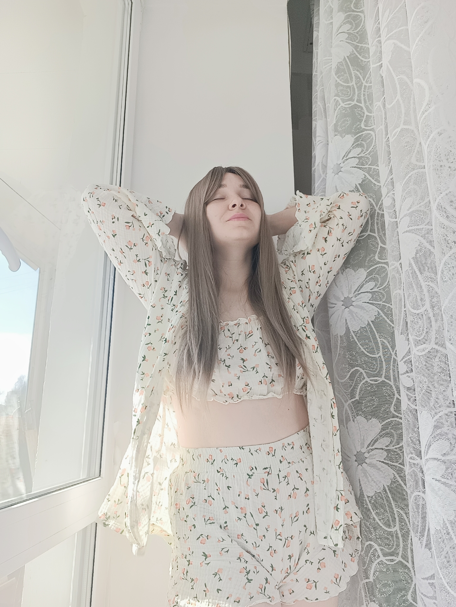 A young woman stands in front of a window with white curtains in an indoor space. She has light brown hair and her eyes are closed. She is wearing a white floral print top and shorts with a matching open-front shirt. She has her hands behind her head. The location is not identifiable as Izhevsk, Russia, based solely on the visual information provided.