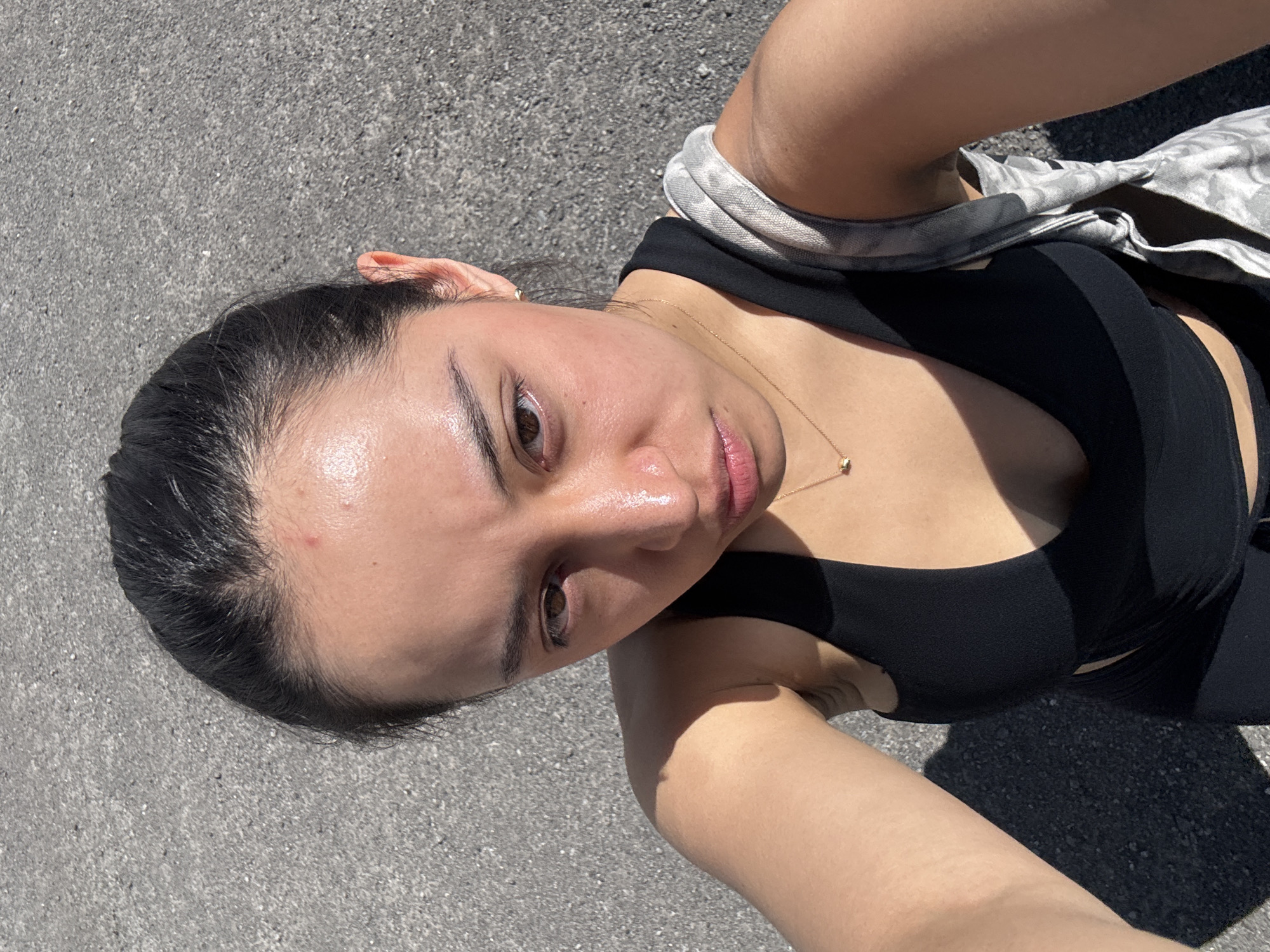 A young woman is captured in a close-up, high-angle selfie taken outdoors in San Bernardino Tlaxcalancingo, Mexico. She is looking directly into the camera lens with a neutral expression, her lips slightly parted. Her dark hair is pulled back, revealing her forehead, which has a natural shine, possibly from sweat or oil. She has dark eyebrows and brown eyes.
She is wearing a simple black tank top or sports bra, and a delicate gold necklace with a small pendant adorns her neck. A light-colored, possibly patterned, strap (potentially from a bag or another garment) is visible over her right shoulder. The background is an expanse of textured, light gray asphalt or concrete, suggesting an outdoor, paved area. The strong, high-contrast lighting and sharp shadows indicate that the photograph was taken during bright daylight hours. No discernible text is visible in the image.