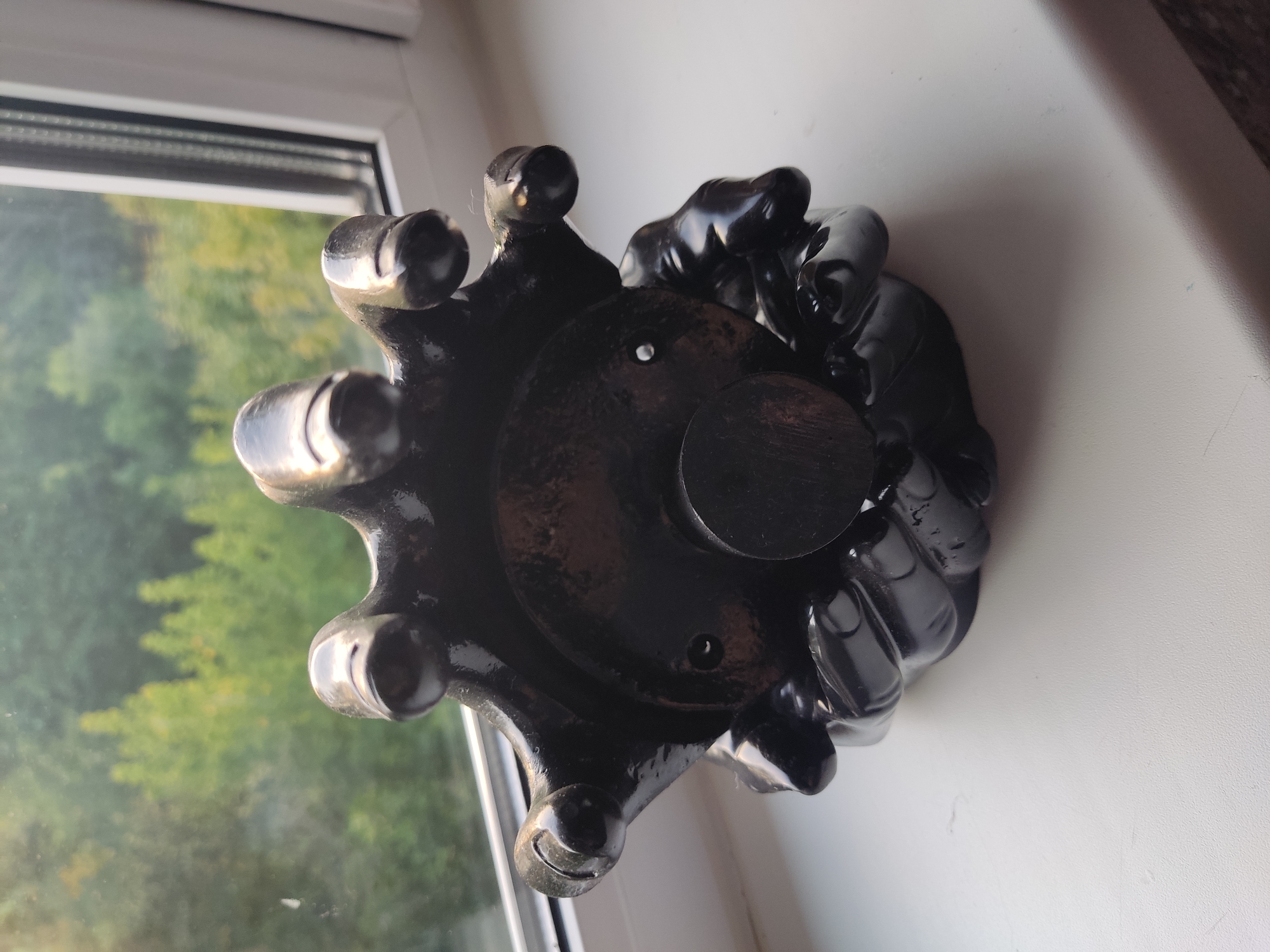 A black, glossy, gothic-style incense burner, shaped like skeletal hands holding a central circular base with a wooden peg, rests on a white windowsill. Beyond the window, lush green trees are visible under a daylight sky. The scene is indoors, with the windowsill acting as a platform. The object appears to be an artifact or decorative item.