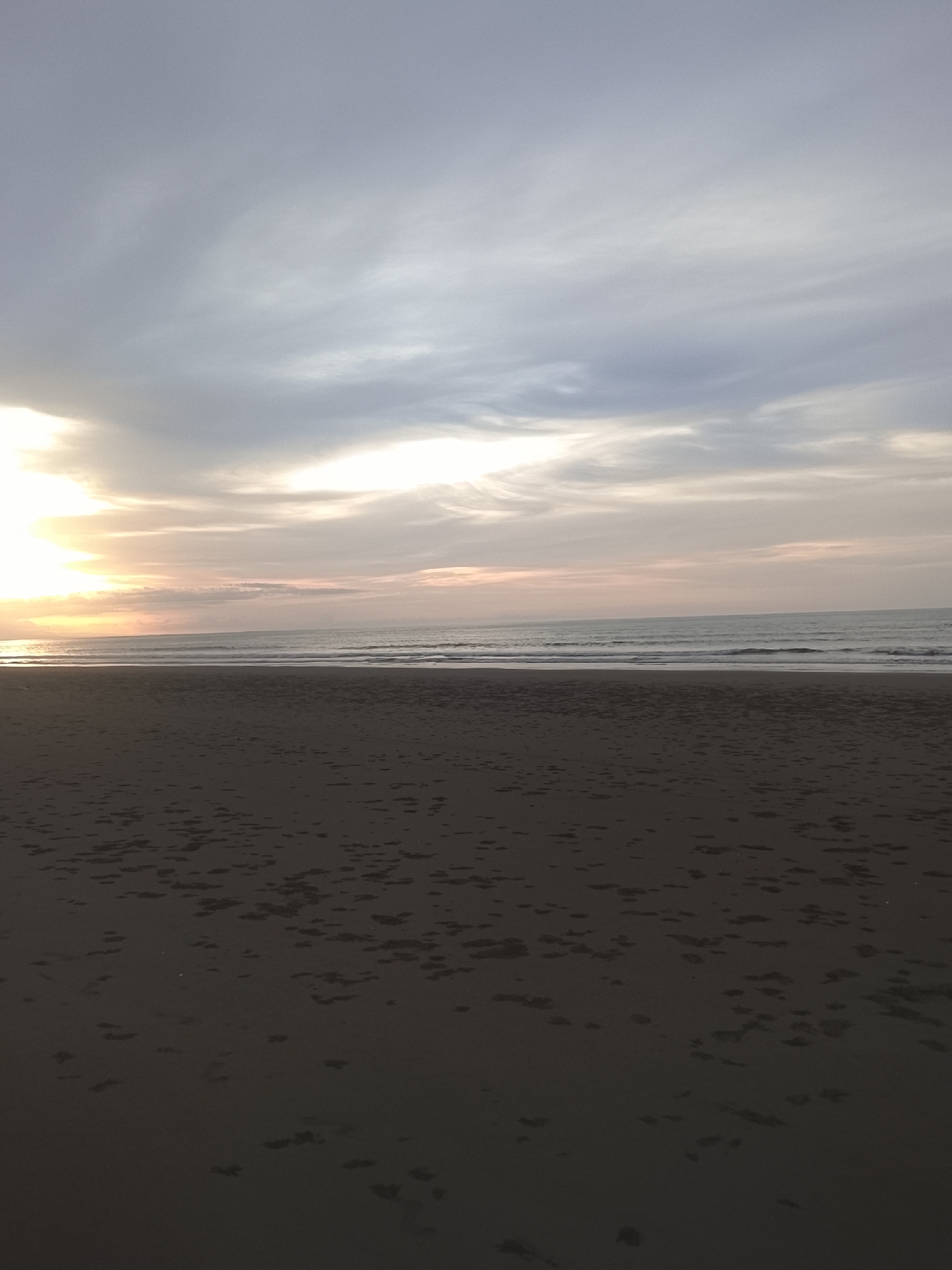 This is a photograph of a beach scene at dusk in Reudeuep, Indonesia. The foreground is dominated by dark, wet sand, marked with numerous footprints and patches of seaweed. The middle ground features a tranquil sea with gentle waves rolling towards the shore. The horizon line is clearly defined, separating the sea from the sky. The sky above is filled with a mix of soft, wispy clouds, with the setting sun casting a warm, golden glow on the left side of the image. The overall atmosphere is serene and peaceful.