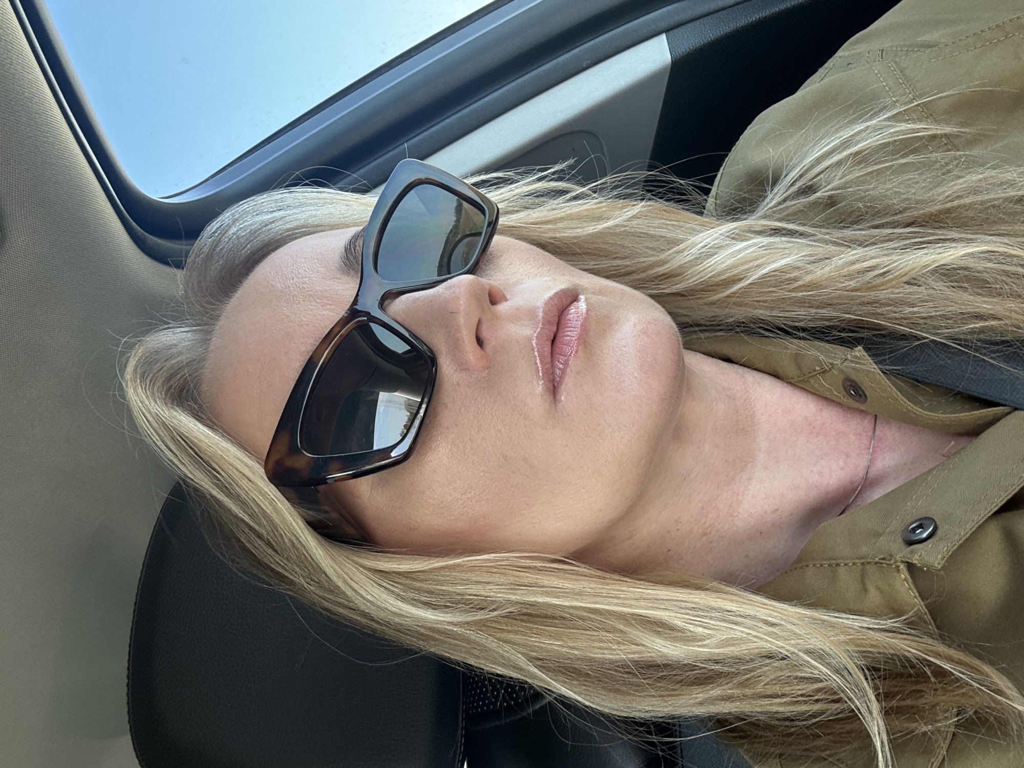 A woman with blonde hair is reclining in the passenger seat of a car. She's wearing sunglasses and an olive green button-down shirt. The car's sunroof is open, revealing a bright blue sky. The location is not discernible as Гавриловка, Russia.