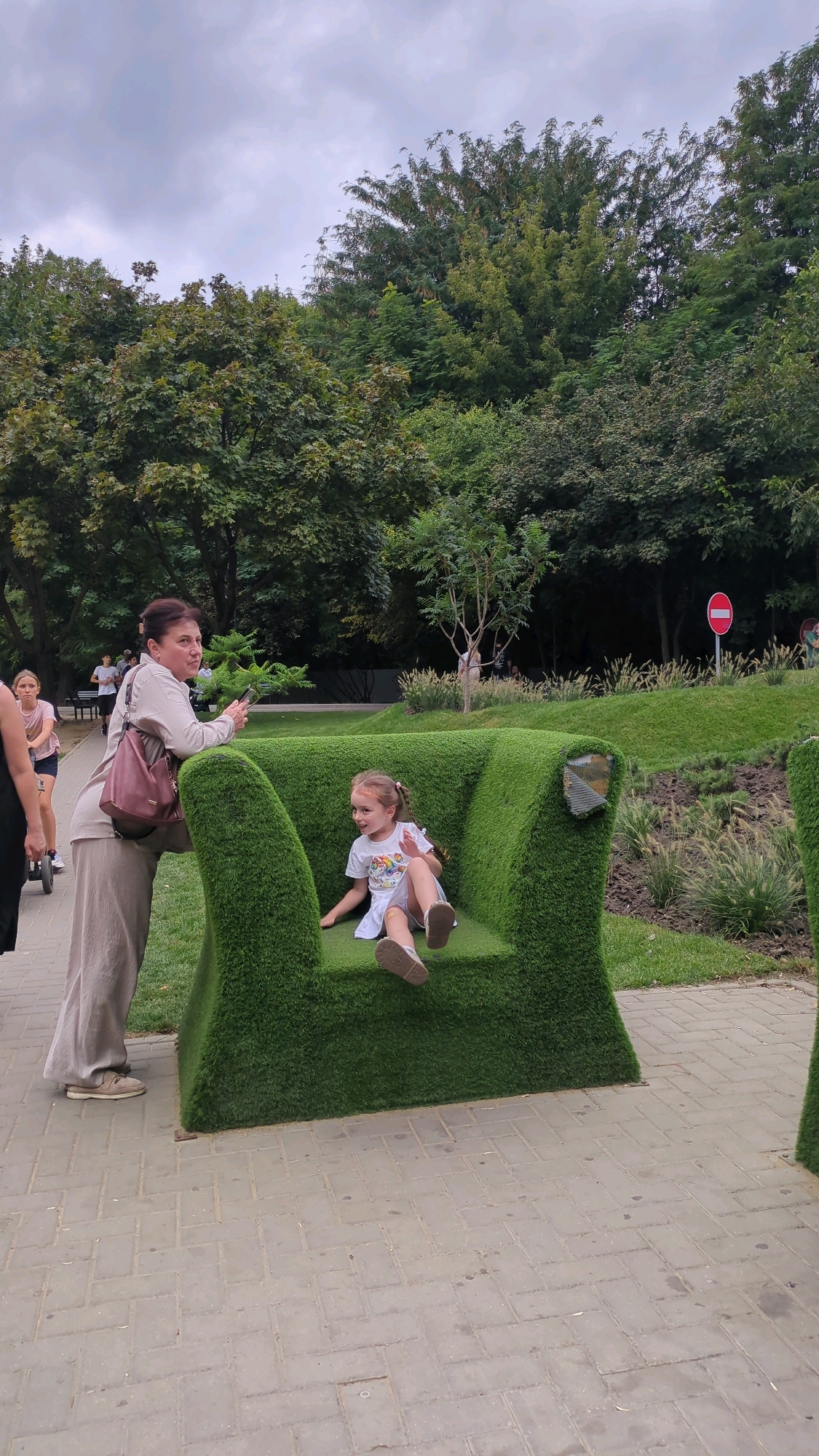 A woman stands next to a large, grass-covered armchair-shaped sculpture in a park, her hand on the back of the sculpture. A young girl with blonde pigtails sits in the grass sculpture, wearing a white t-shirt and shorts. The park has trees and a paved walkway in the foreground. A red and white circular sign is visible in the background on a pole. The scene appears to be in Chișinău, Moldova, given the context provided.