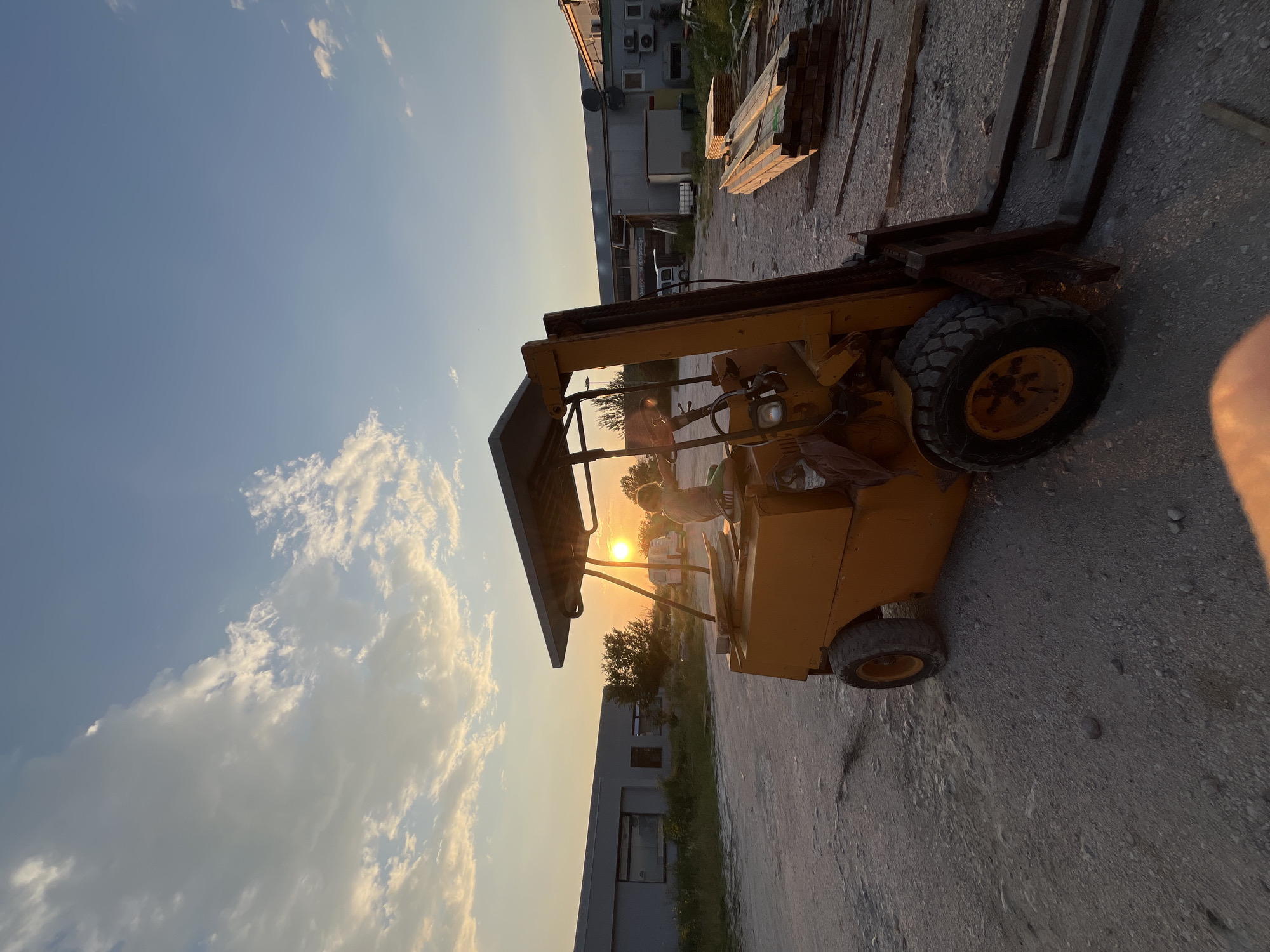 The image shows a yellow forklift parked on a paved surface in what appears to be an industrial or construction area in Pobrdje, Montenegro. The sun is setting, casting a warm glow across the scene. A child is seated in the forklift's driver seat. In the background, there are buildings, trees, and stacks of construction materials. The sky is visible with some clouds.
