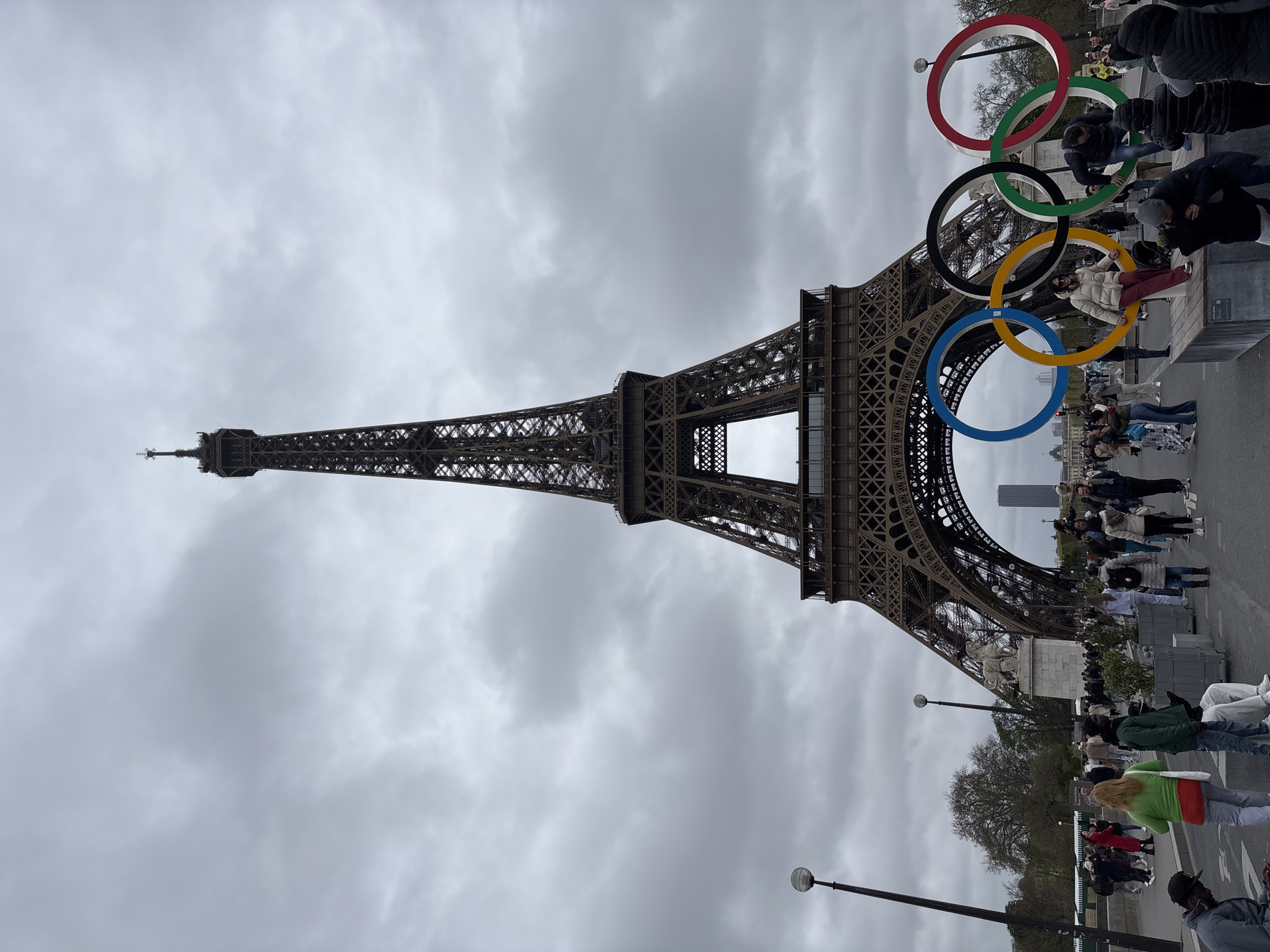 The image captures the Eiffel Tower in Paris, France, against a cloudy sky. In the foreground, the Olympic rings are displayed, with tourists interacting with them. There are people posing for photos and walking around, creating a lively tourist scene at the base of the tower. Street lamps are visible.