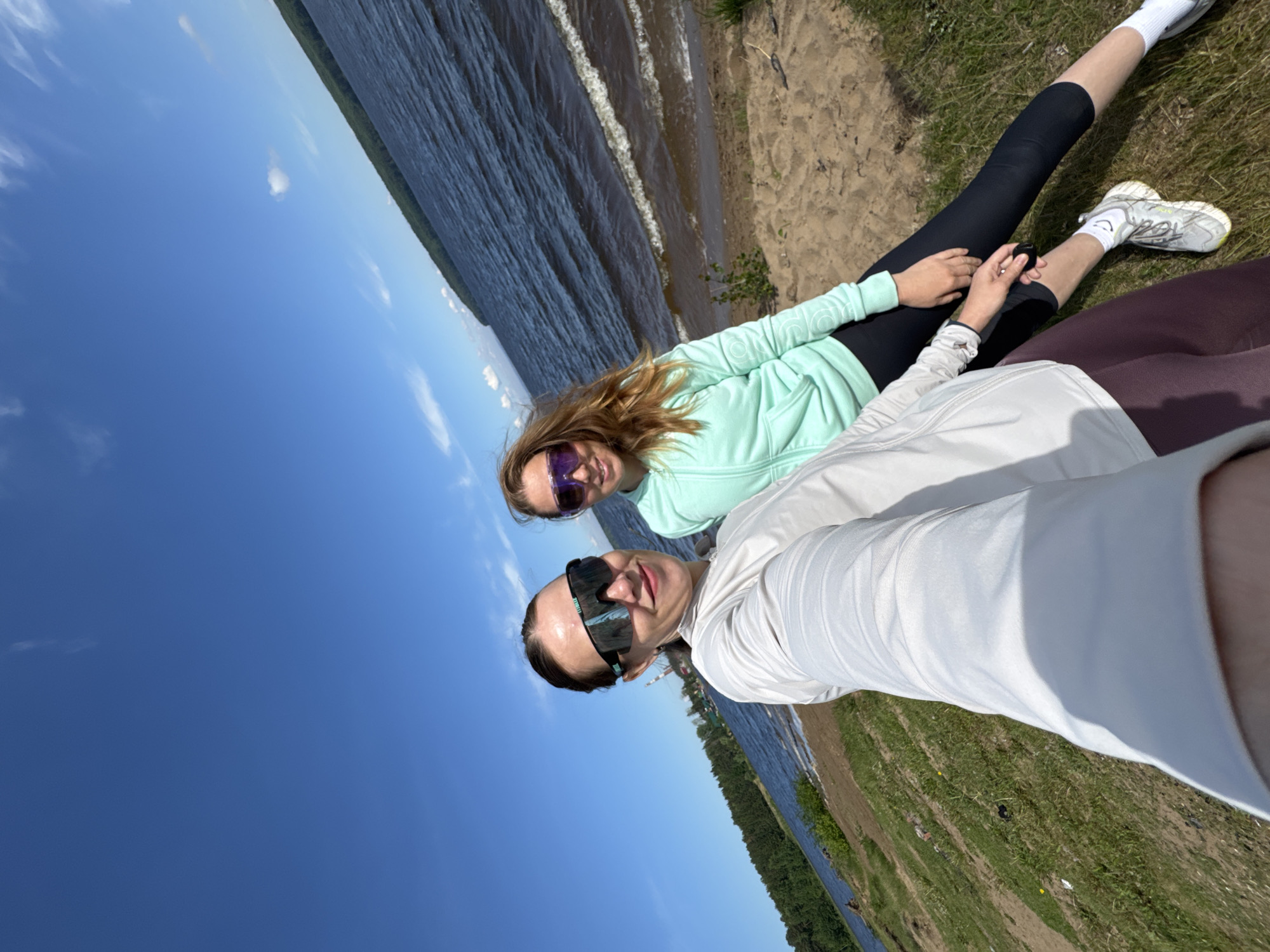 The image depicts a selfie taken by two young women enjoying a sunny day outdoors in Chaikovsky, Russia.
One woman, likely taking the photo, is in the immediate foreground, wearing a white long-sleeved top and dark, futuristic-style sunglasses. She has dark hair pulled back and looks towards the camera with a neutral or slightly smiling expression. Behind her, a second woman with long, reddish-brown hair, wearing a mint green hooded top, dark leggings, white socks, and light-colored sneakers, sits with her hands clasped. She also wears reflective purple sunglasses and smiles towards the camera.
They are positioned on a sandy and grassy shoreline beside a large body of dark blue water, which shows gentle waves. The background features a clear blue sky dotted with scattered small white clouds, and a distant green treeline on the opposite bank of the water body. The bright sunlight suggests a daytime setting, possibly late morning or afternoon. The overall atmosphere conveys relaxation and enjoyment of the natural surroundings. No visible text is present in the image.